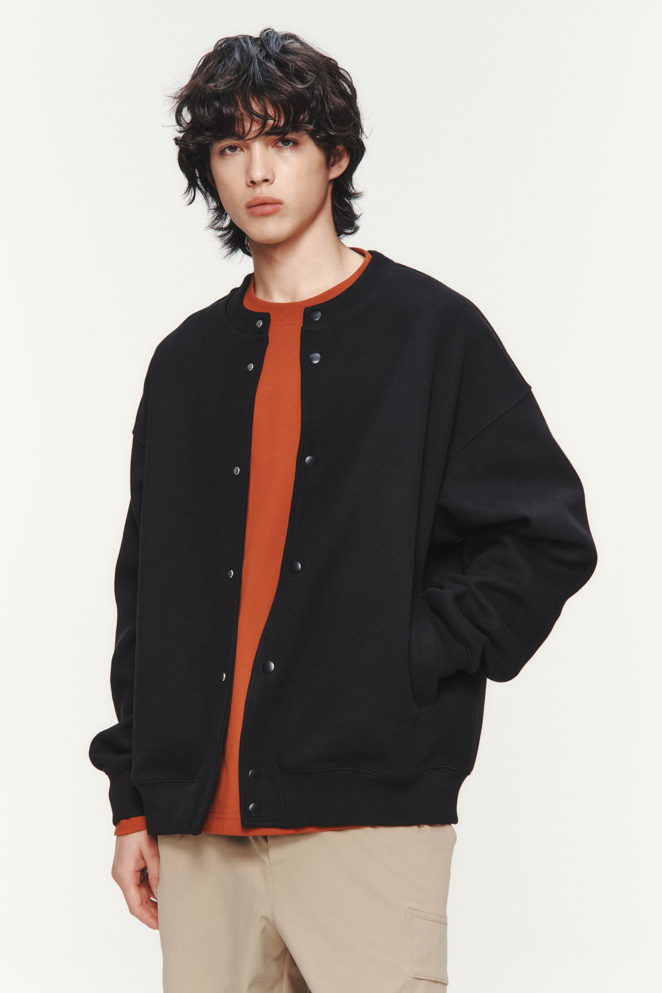 이미지 확대 보기: 모델이 착용한 블랙 와플 가디건은 둥근 리브 트림 넥라인, 드롭 숄더, 넓은 리브 커프스가 있는 긴 소매, 넓은 리브 헴이 특징입니다. 가디건은 앞면에 프레스 스터드가 있으며, 일부는 풀려 있고, 웰트 프론트 포켓이 있어 모델의 오른손이 그 중 하나에 들어가 있으며, 오렌지색 티셔츠 위에 레이어드 되어 있습니다.