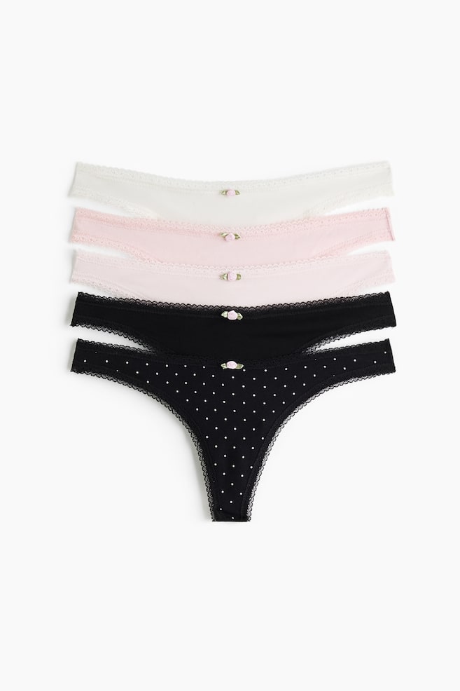 Cinco pares de cuecas tanga de cintura baixa são apresentados de frente numa disposição empilhada, exibindo modelos em branco, rosa-claro, rosa-pálido, preto liso e preto às bolinhas. Cada par apresenta uma delicada aplicação de renda ao longo das bordas e um pequeno detalhe de botão de rosa em tecido no centro da cintura. - 1