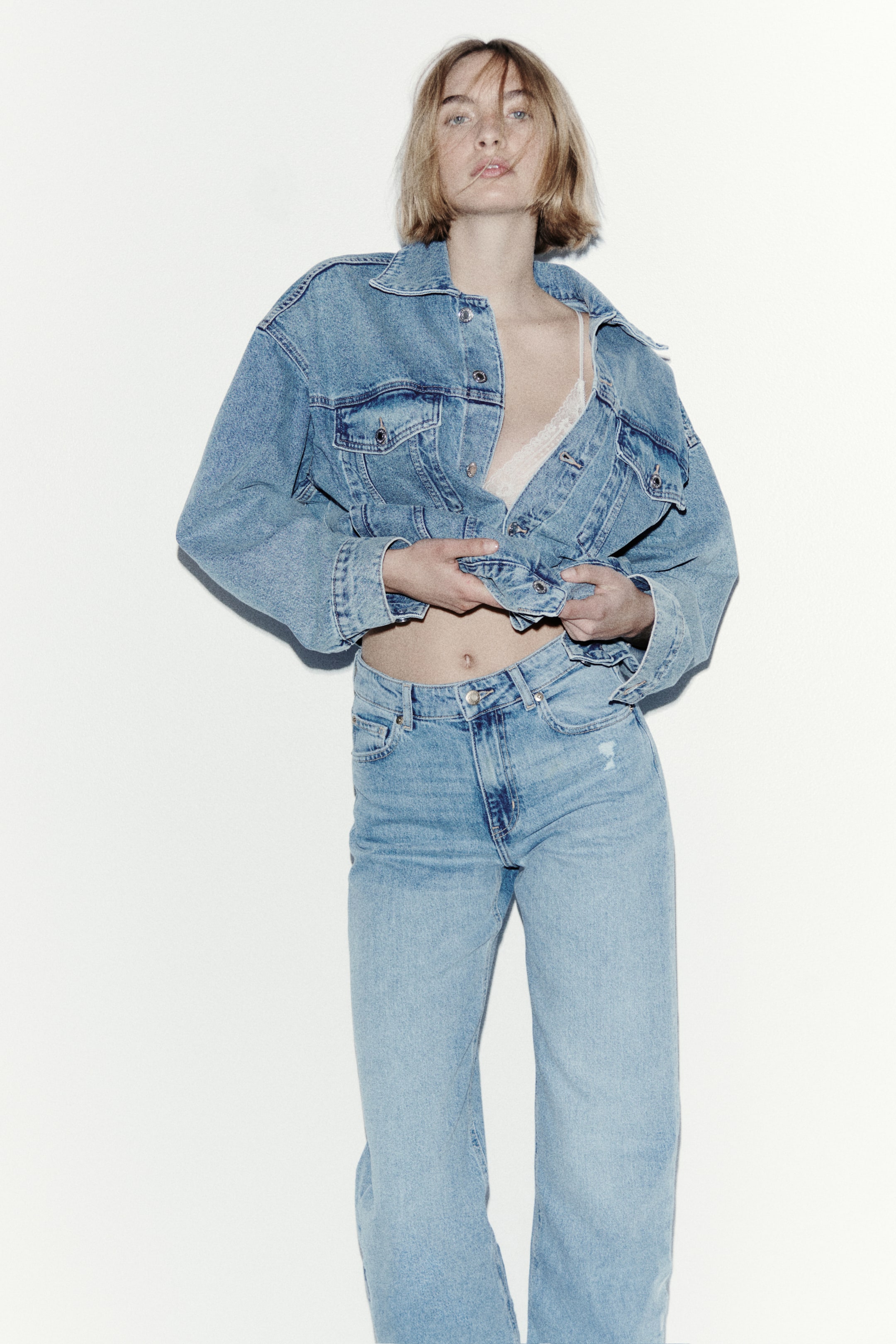 Vezi imaginea mai mare: O jachetă trucker din denim spălat deschis, supradimensionată, cu un guler ascuțit, buzunare la piept cu clapă și nasturi și mâneci lungi cu manșete cu nasturi, având partea din față deschisă pentru a dezvălui un bralette din dantelă. Jacheta este completată de o pereche de blugi cu talie înaltă, croială dreaptă, din denim spălat deschis, care prezintă detalii uzate pe coapse.