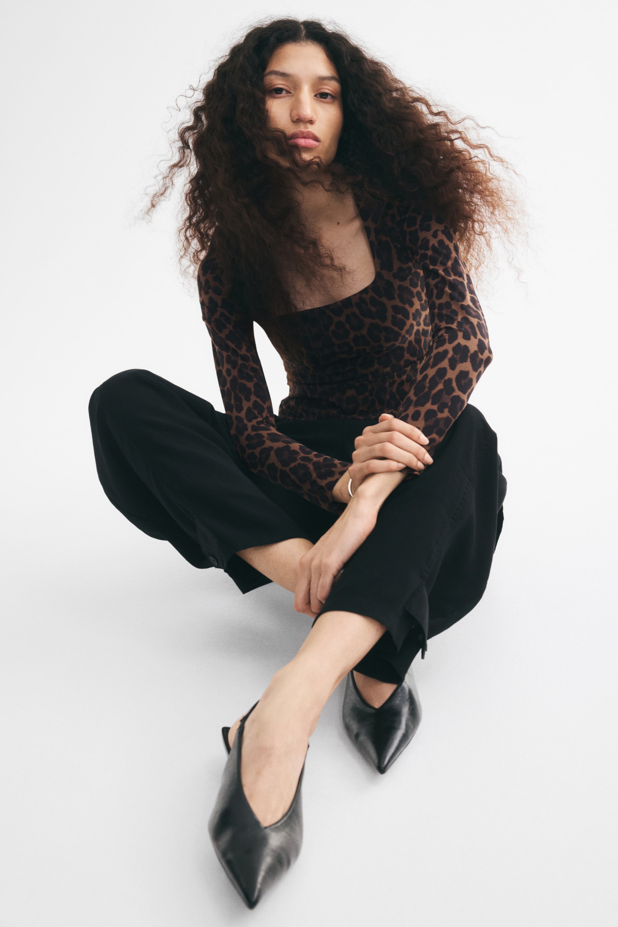 View larger image: A fitted leopard-print top with a deep square neckline and long sleeves is worn by the model. The top is styled with wide-leg black trousers and black pointed-toe shoes as the model sits with her legs crossed.