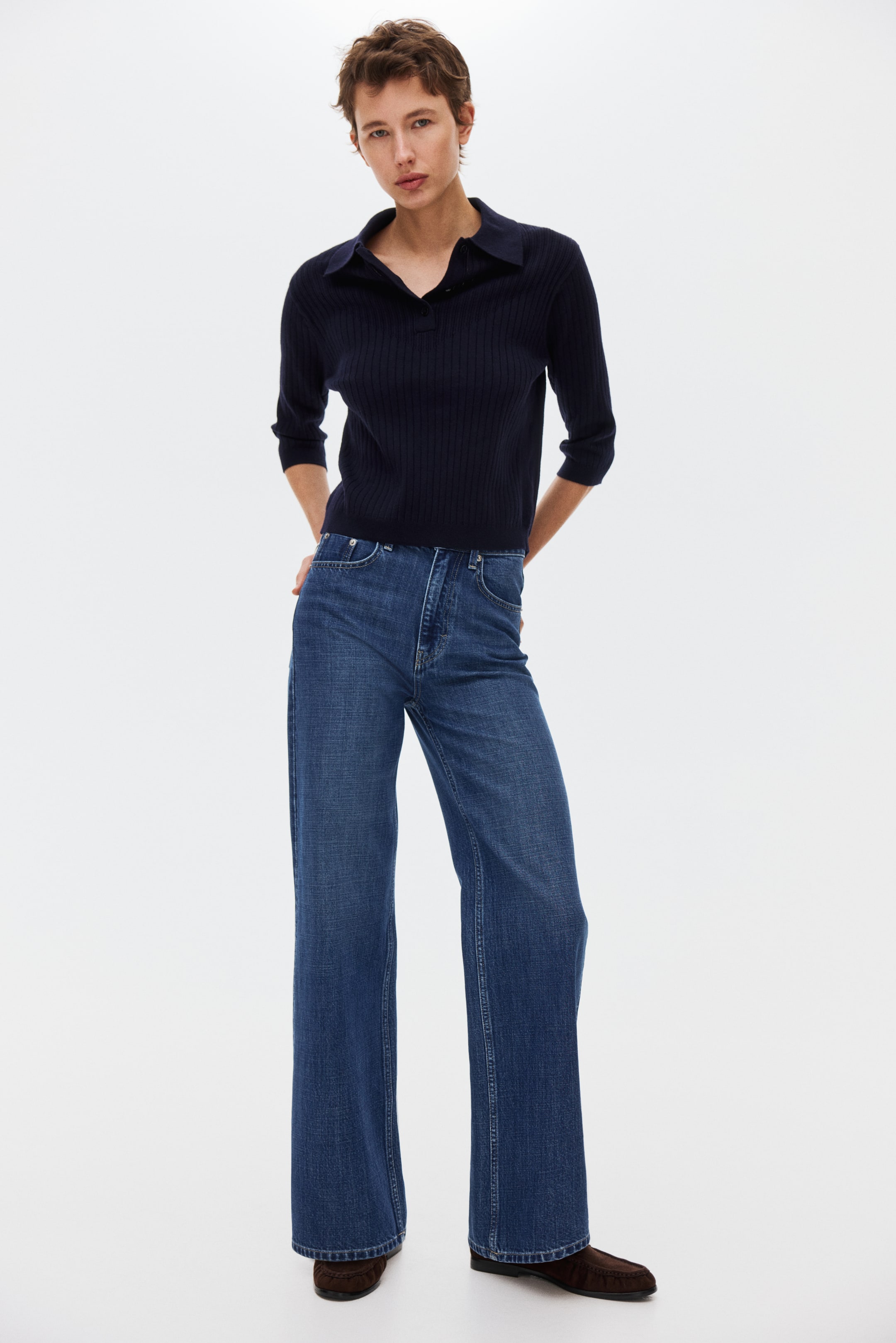 Agrandir l'image: Jeans en denim bleu foncé présentant une silhouette taille haute à jambe large. Ils arborent une coupe classique à cinq poches, des surpiqûres apparentes et une fermeture avant par bouton et fermeture à glissière.