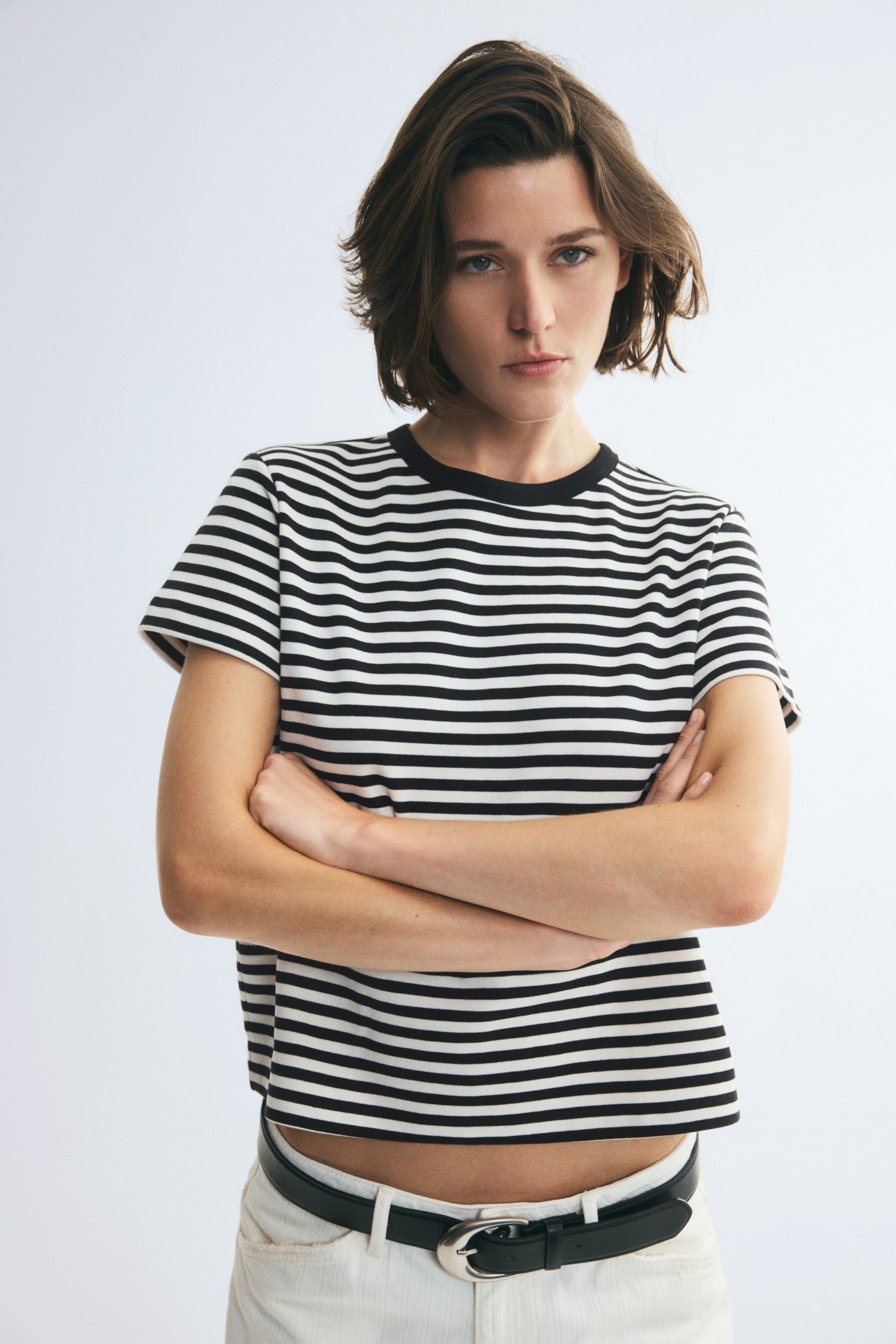 View larger image: A fitted, short-sleeved T-shirt with horizontal black-and-white stripes and a solid black ribbed crew neckline is worn by the model. The tee features a straight-cut hem and is styled with white trousers and a black belt.