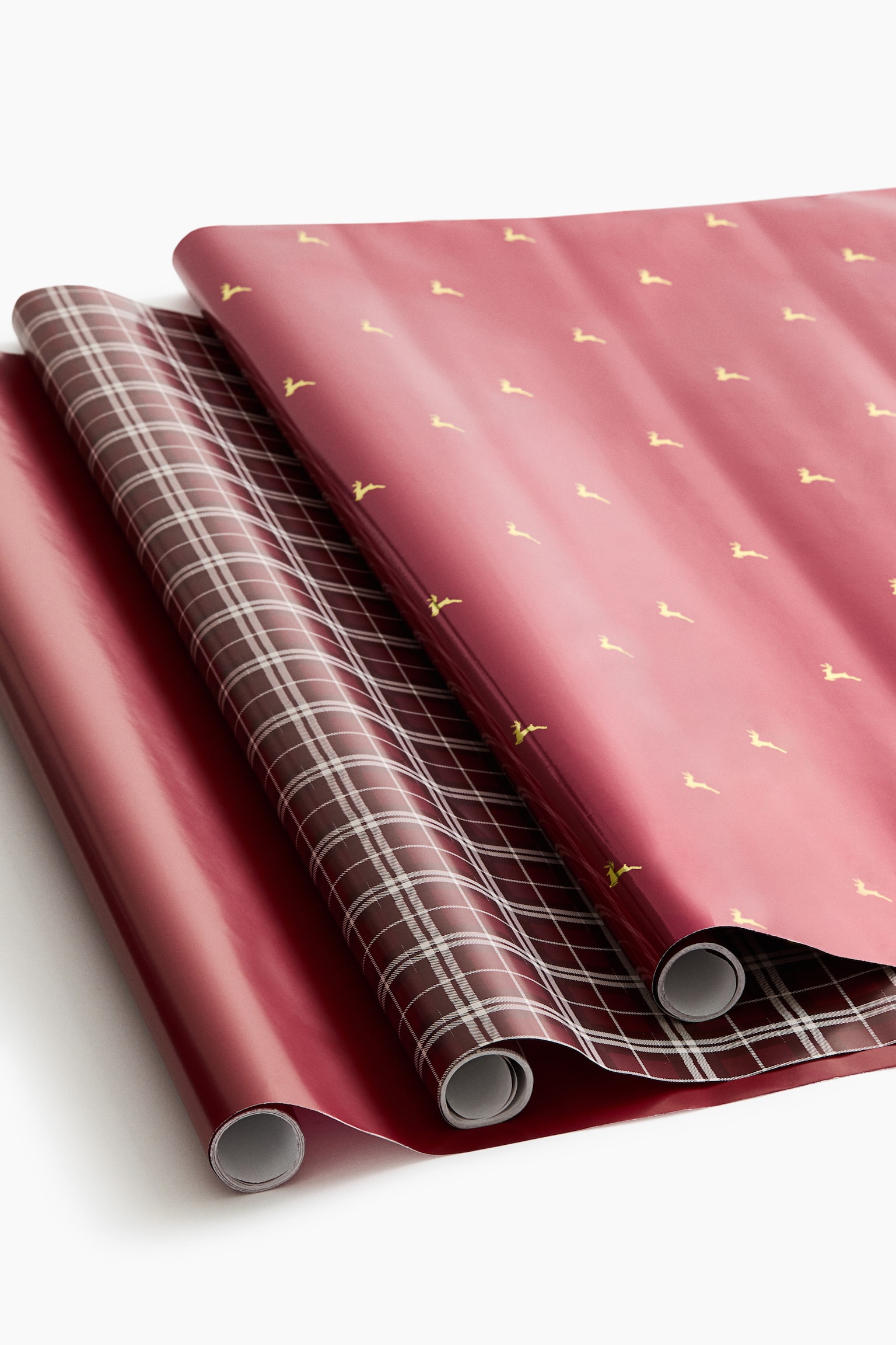 Trois rouleaux de papier cadeau sont partiellement dÃ©roulÃ©s et superposÃ©s, mettant en valeur leurs motifs coordonnÃ©s. Le rouleau du dessus prÃ©sente un fond rouge ornÃ© de petites silhouettes de rennes dorÃ©es, le rouleau du milieu affiche un motif Ã  carreaux bordeaux et blanc, et le rouleau du dessous est uni, avec une finition bordeaux brillante.