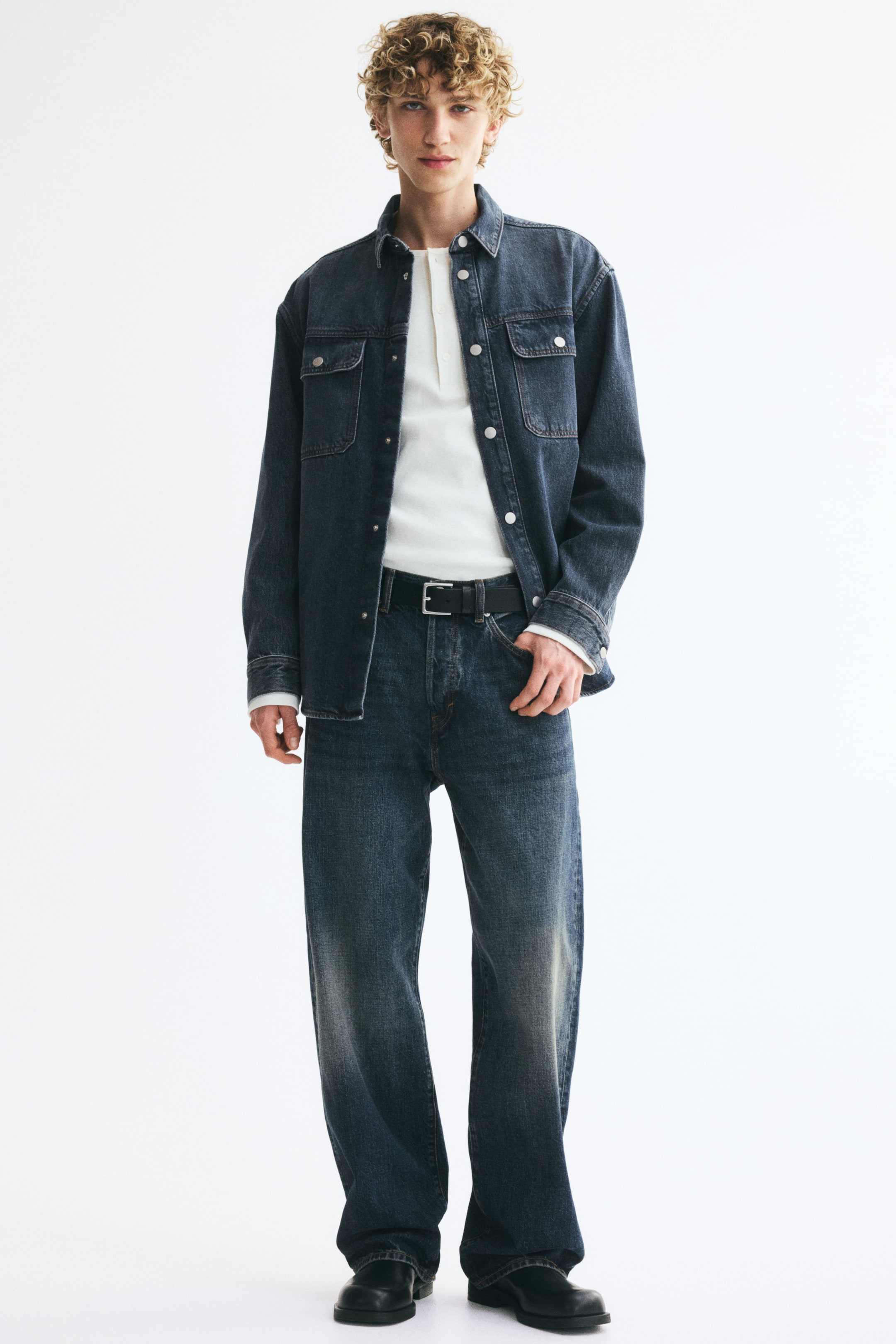 Xem ảnh lớn hơn: Quần jeans 5 túi bằng denim cotton cứng màu xanh đậm, với cạp vừa, khóa kéo, đáy thụng và ống quần bo tròn dáng rộng từ phần mông đến gấu, tạo thêm độ thoải mái, đang được người mẫu mặc. Người mẫu kết hợp quần jeans với thắt lưng màu đen, một tay đang đặt trong túi trước.