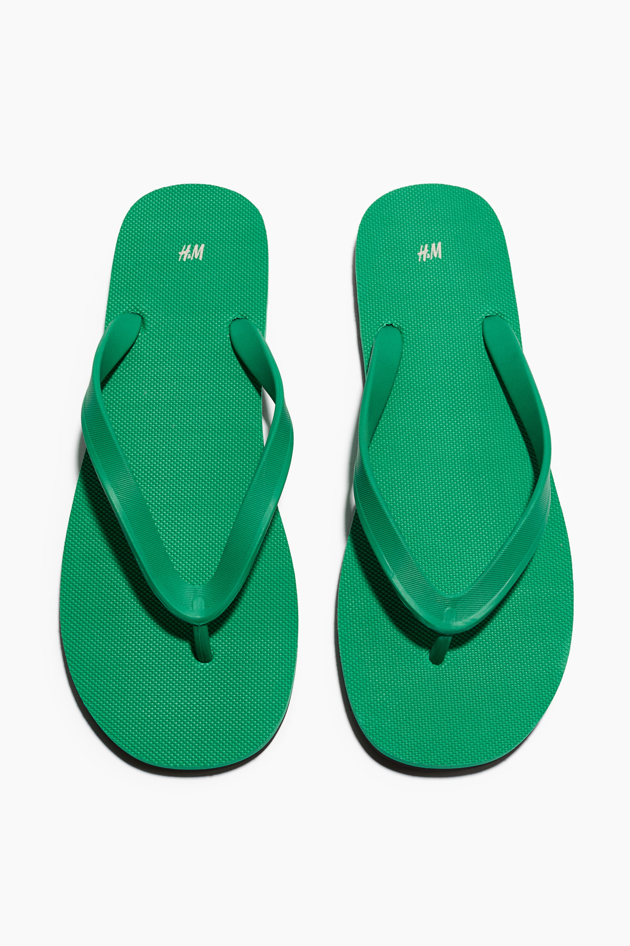 Grotere afbeelding bekijken: Twee groene rubberen teenslippers met een getextureerd voetbed en gladde V-vormige bandjes die aansluiten op een smalle teenpost. Witte "H.M" branding is zichtbaar op de hiel van elke teenslipper.