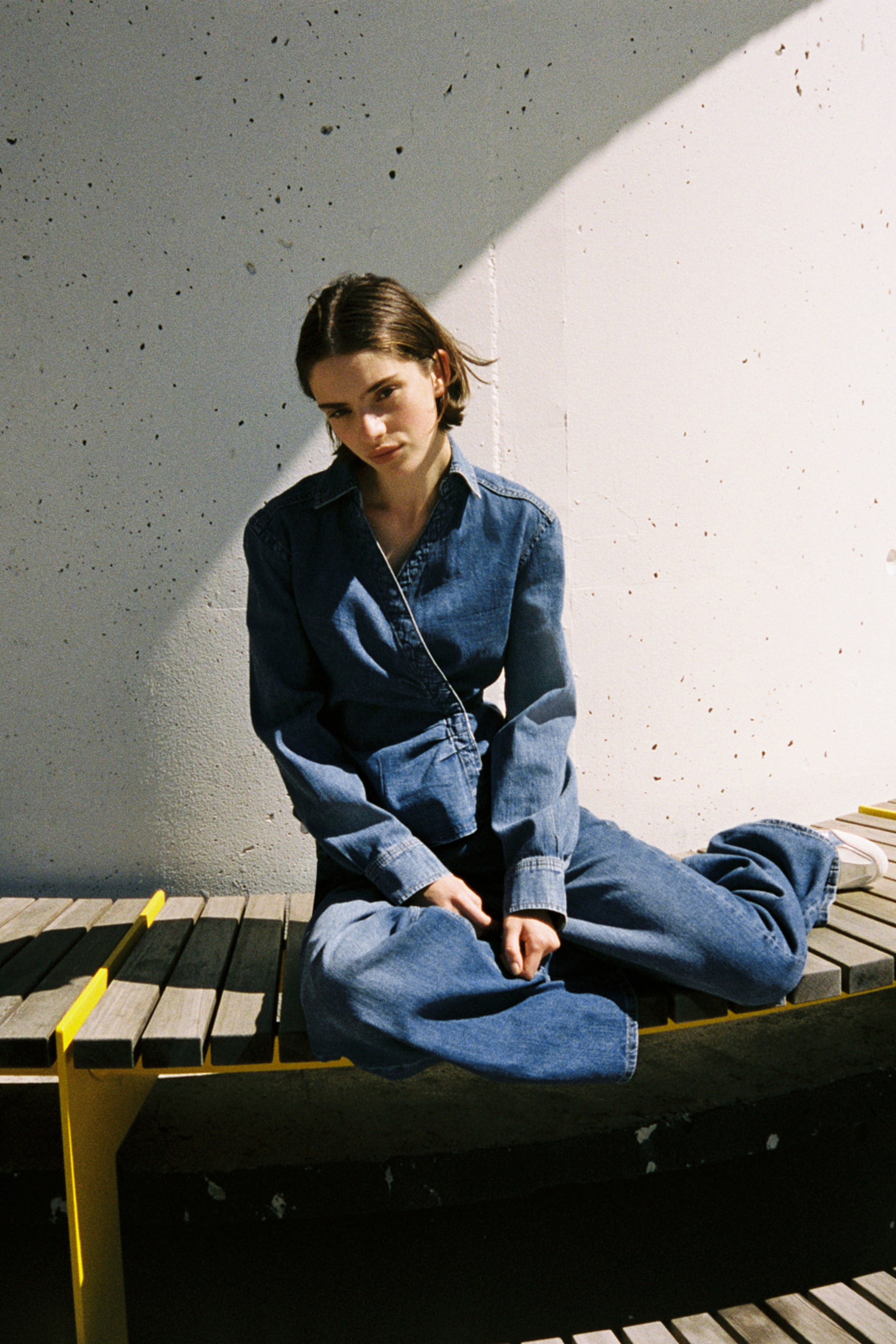 Voir une image plus grande: La mannequin porte une chemise en denim ajustée, avec un col ouvert, un devant croisé avec des fronces créant un effet drapé, des manches longues avec poignets boutonnés, un empiècement au dos et un ourlet légèrement arrondi. La mannequin est assise, complétant la tenue avec un pantalon en denim assorti.