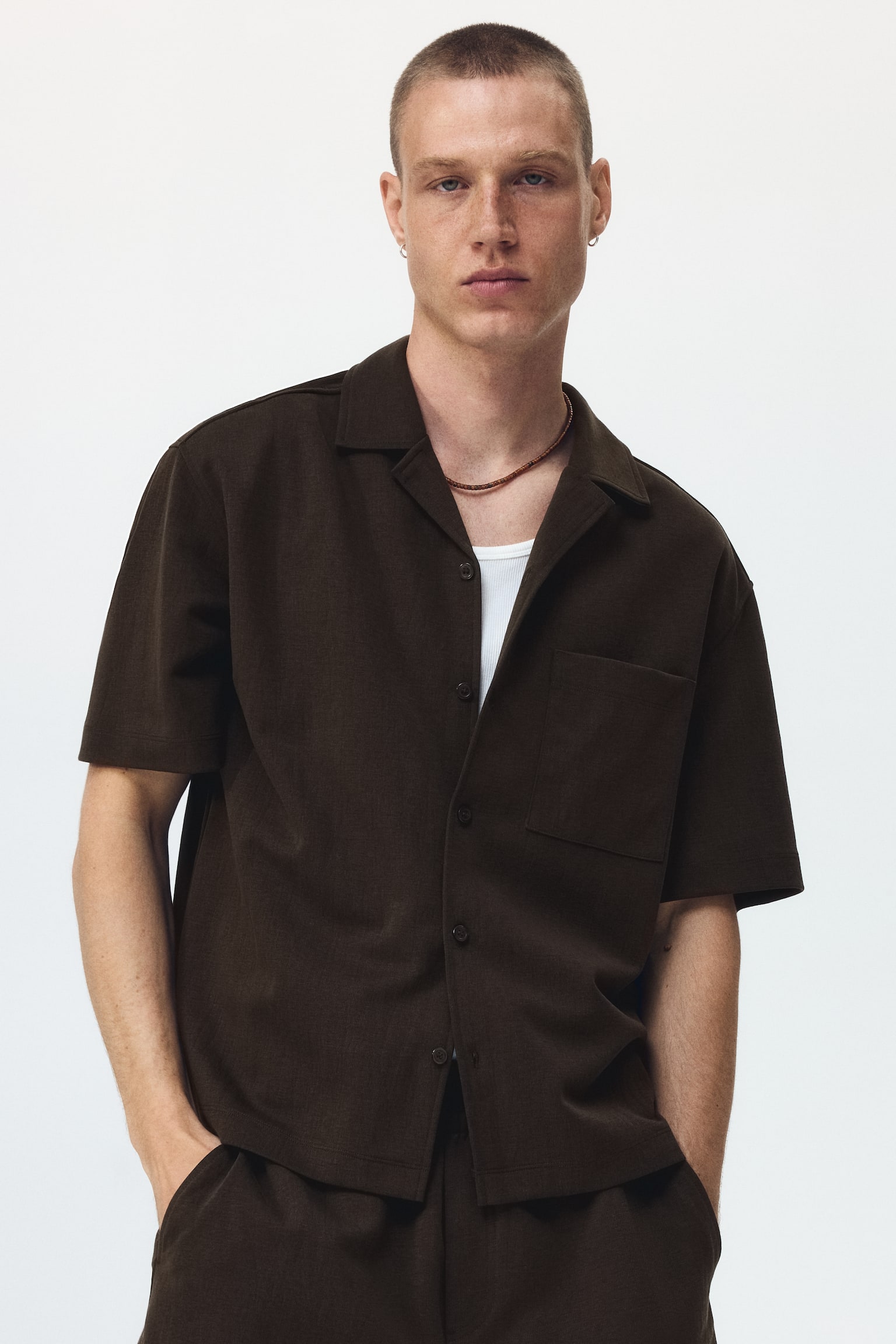 A dark brown short-sleeved shirt is facing front, featuring a camp collar and a full button-front closure with dark buttons. It is displaying a single patch pocket on the left chest, short sleeves, and a subtly textured fabric. - 1