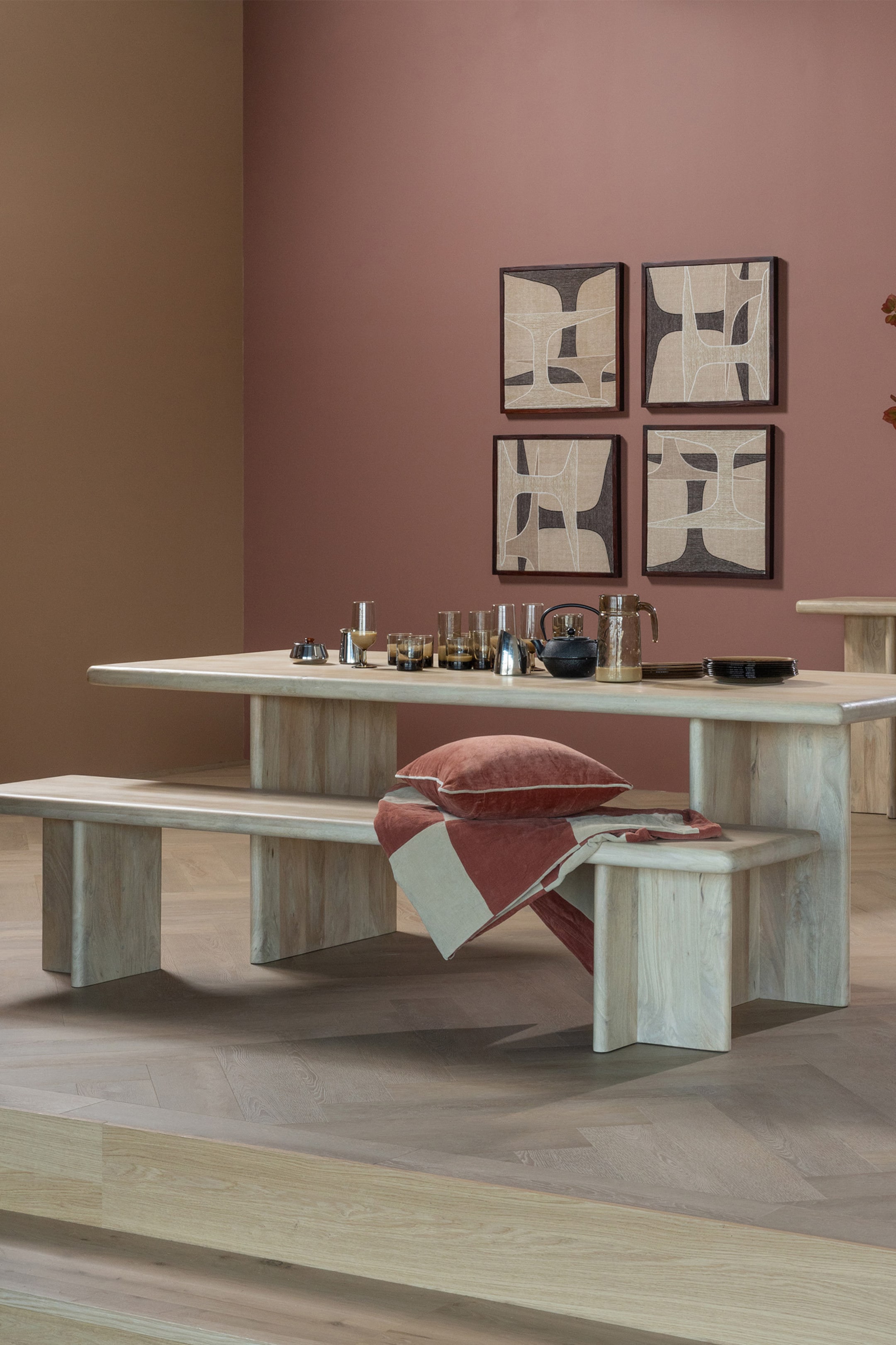 Agrandir l'image: Une table à manger en bois clair et deux bancs assortis sont présentés sous un léger angle, mettant en valeur leurs épais plateaux et assises rectangulaires, chacun soutenu par deux pieds rectangulaires épais. La table accueille divers articles en verre, une théière et de petits plats, tandis qu'un banc présente un coussin en velours couleur rouille et une couverture rayée drapée dessus.