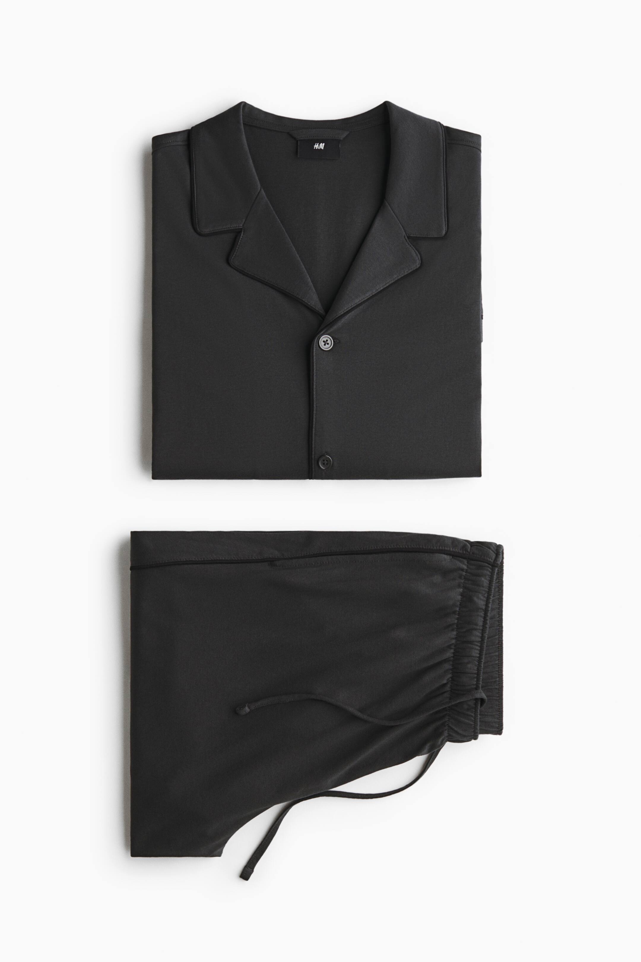 View larger image: A black pyjama set is laid flat in a front view, featuring a neatly folded button-up shirt with notch lapels and matching folded trousers with an elastic drawstring waistband.