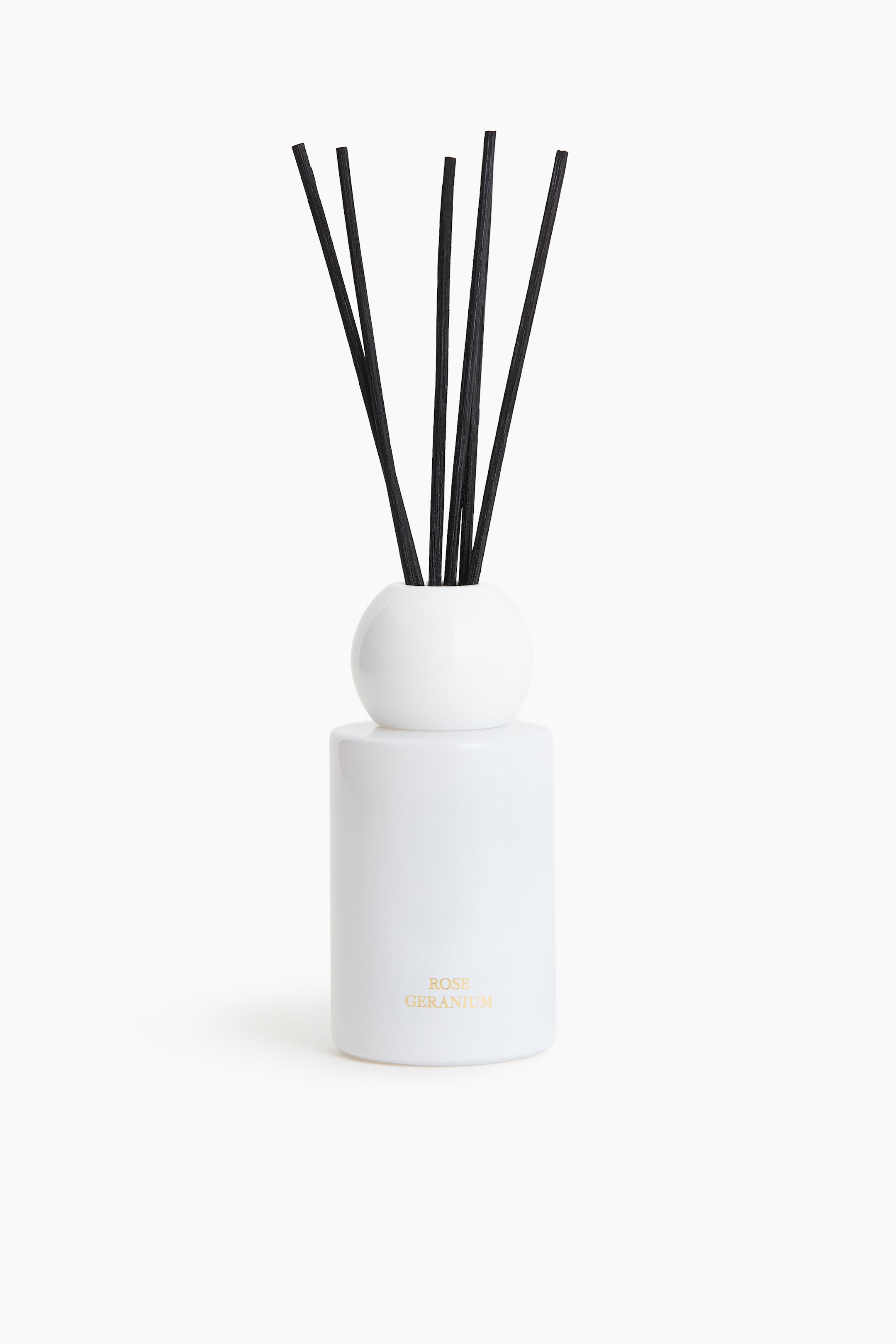 View larger image: An opaque white cylindrical diffuser bottle is standing in the foreground, featuring a spherical top. Several black wooden reeds are emerging from the top, and gold text reading 'ROSE GERANIUM' is visible on the front of the bottle.
