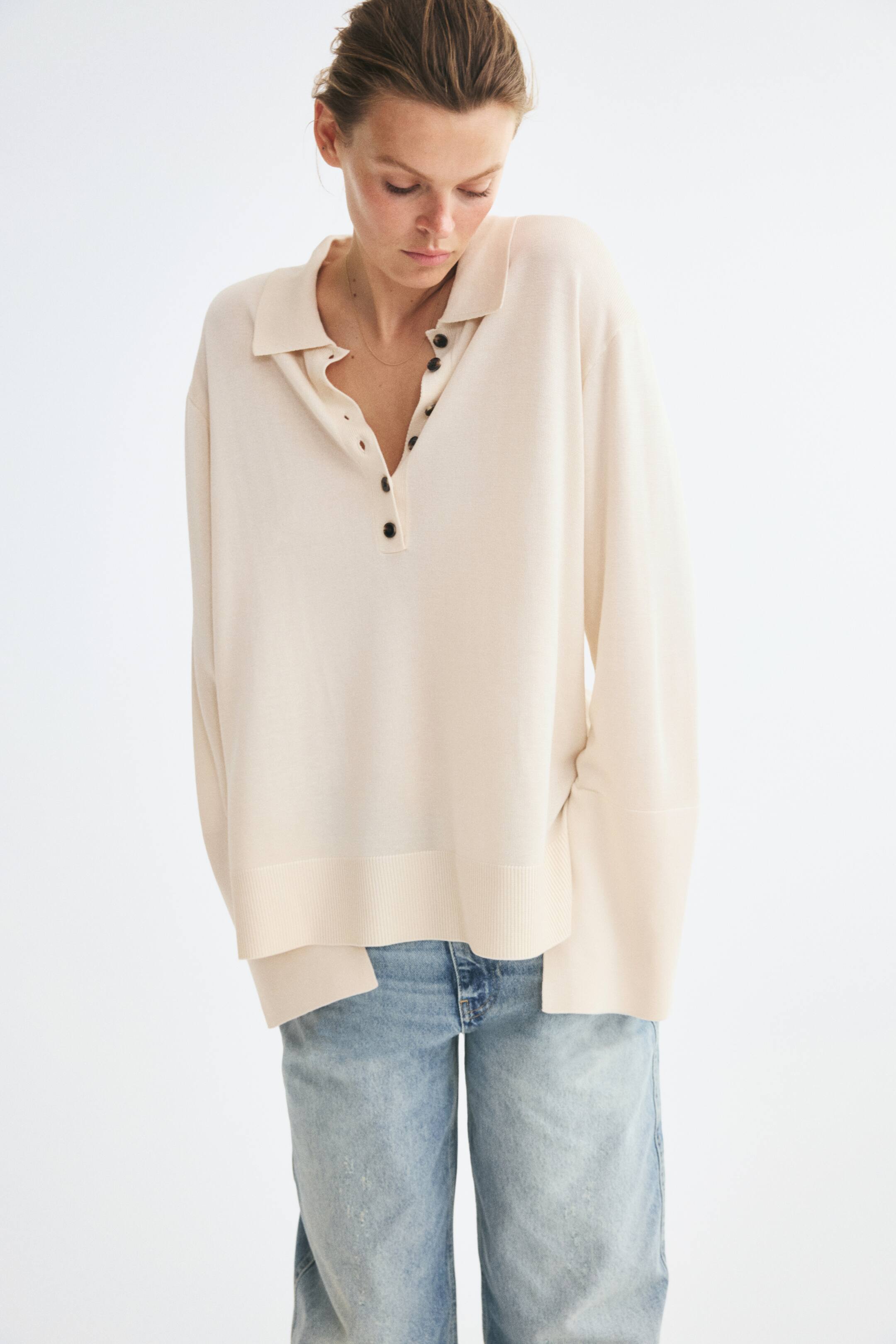 View larger image: The model is wearing a cream-colored, fine-knit polo shirt featuring a ribbed collar, an open button placket with dark buttons, dropped shoulders, and long, wide sleeves with split cuffs. The relaxed-fit shirt is paired with light-wash denim jeans.