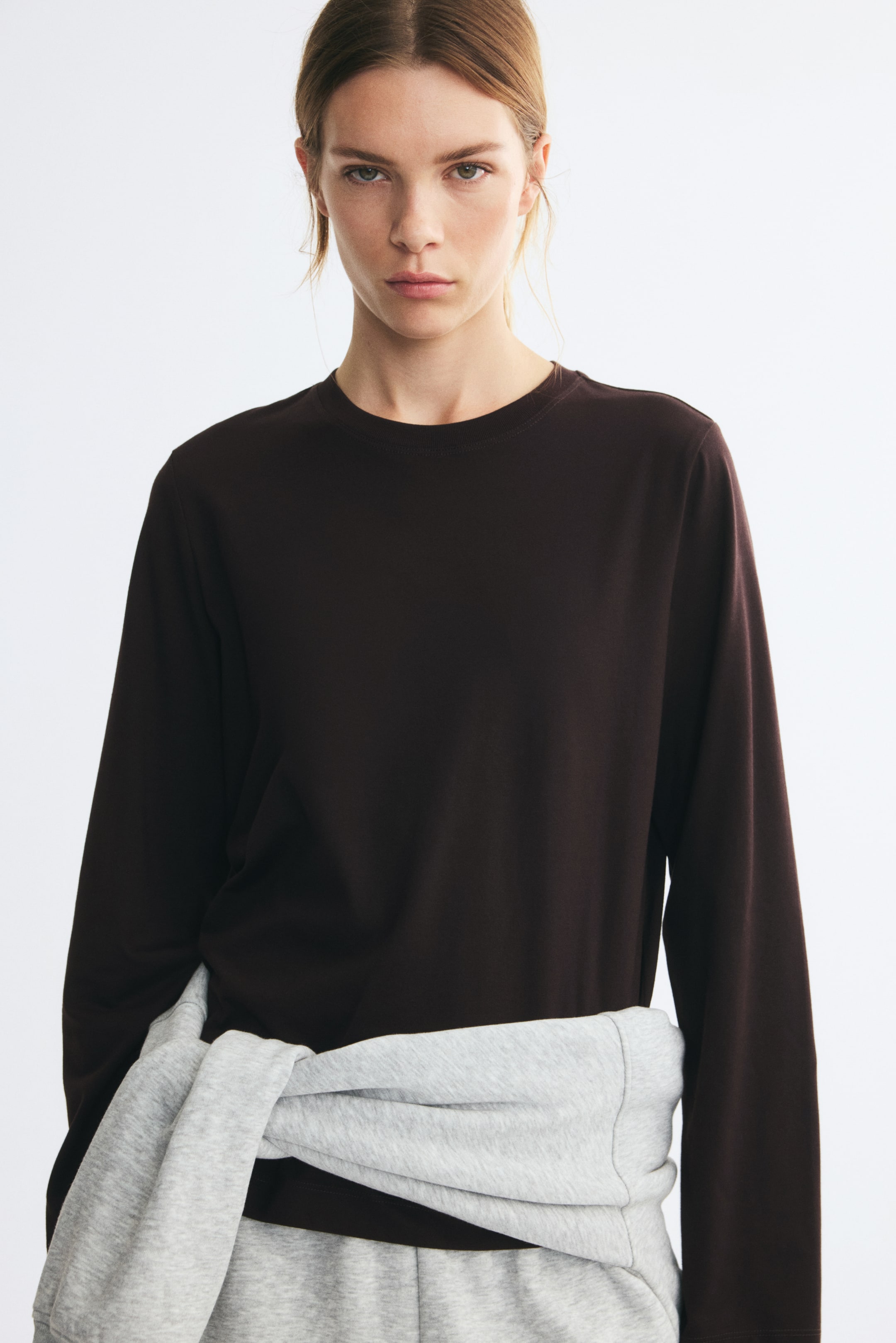 Größeres Bild ansehen: Ein dunkelbraunes Langarm-T-Shirt mit Rundhalsausschnitt wird vom Model getragen, dazu ein hellgrauer Sweatshirt, das um die Taille gebunden ist.
