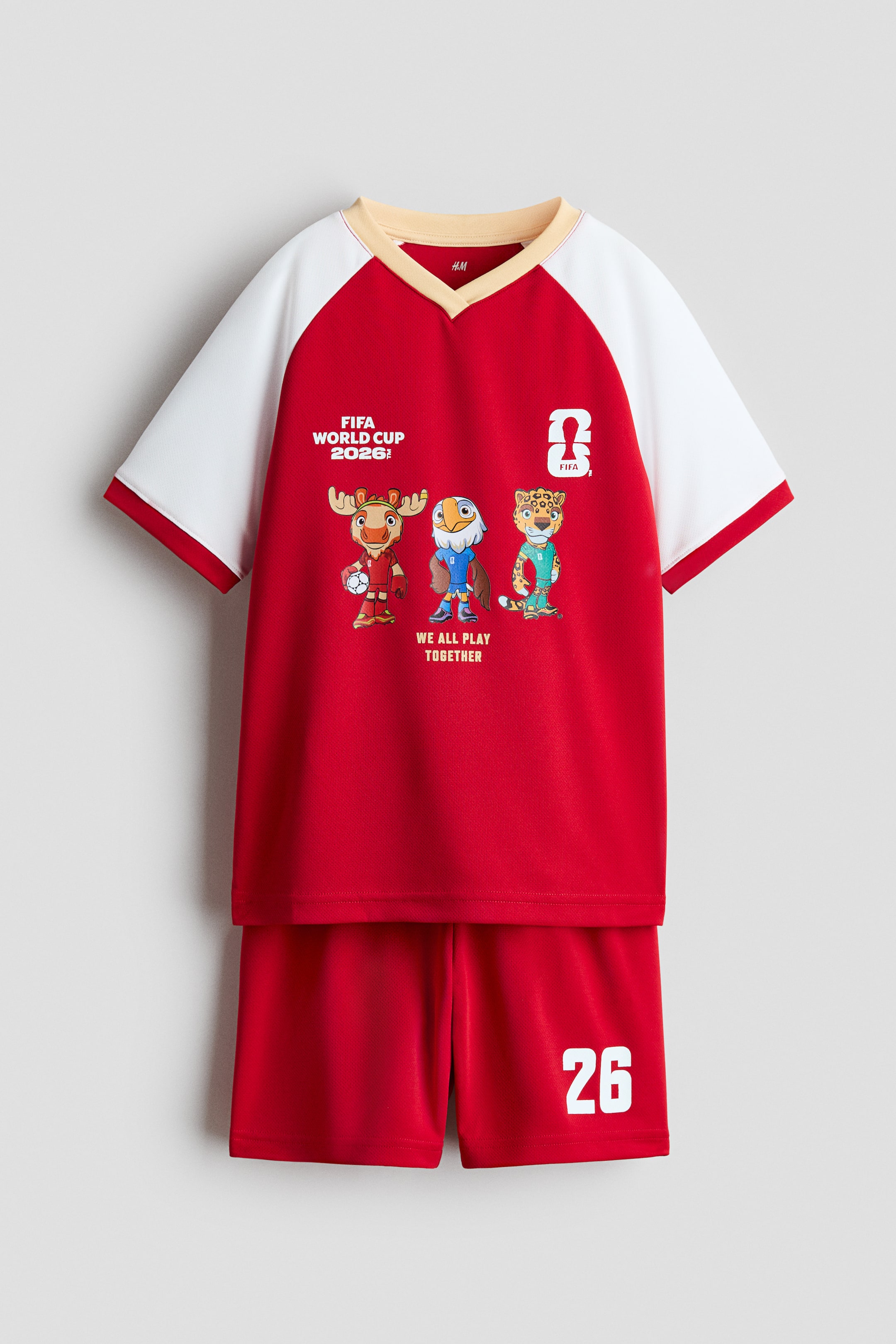 Größeres Bild ansehen: Ein Fußballtrikot und -shorts-Set wird von vorne präsentiert. Es zeigt ein rotes Trikot mit weißen Raglanärmeln und einem beige gerippten V-Ausschnitt. Auf dem Trikot steht "FIFA WORLD CUP 2026!", eine stilisierte "2" mit "FIFA", drei Cartoon-Tiermaskottchen und "WE ALL PLAY TOGETHER" auf der Vorderseite. Die passenden roten Shorts haben einen elastischen Bund und die weiße Nummer "26" auf dem rechten Bein.