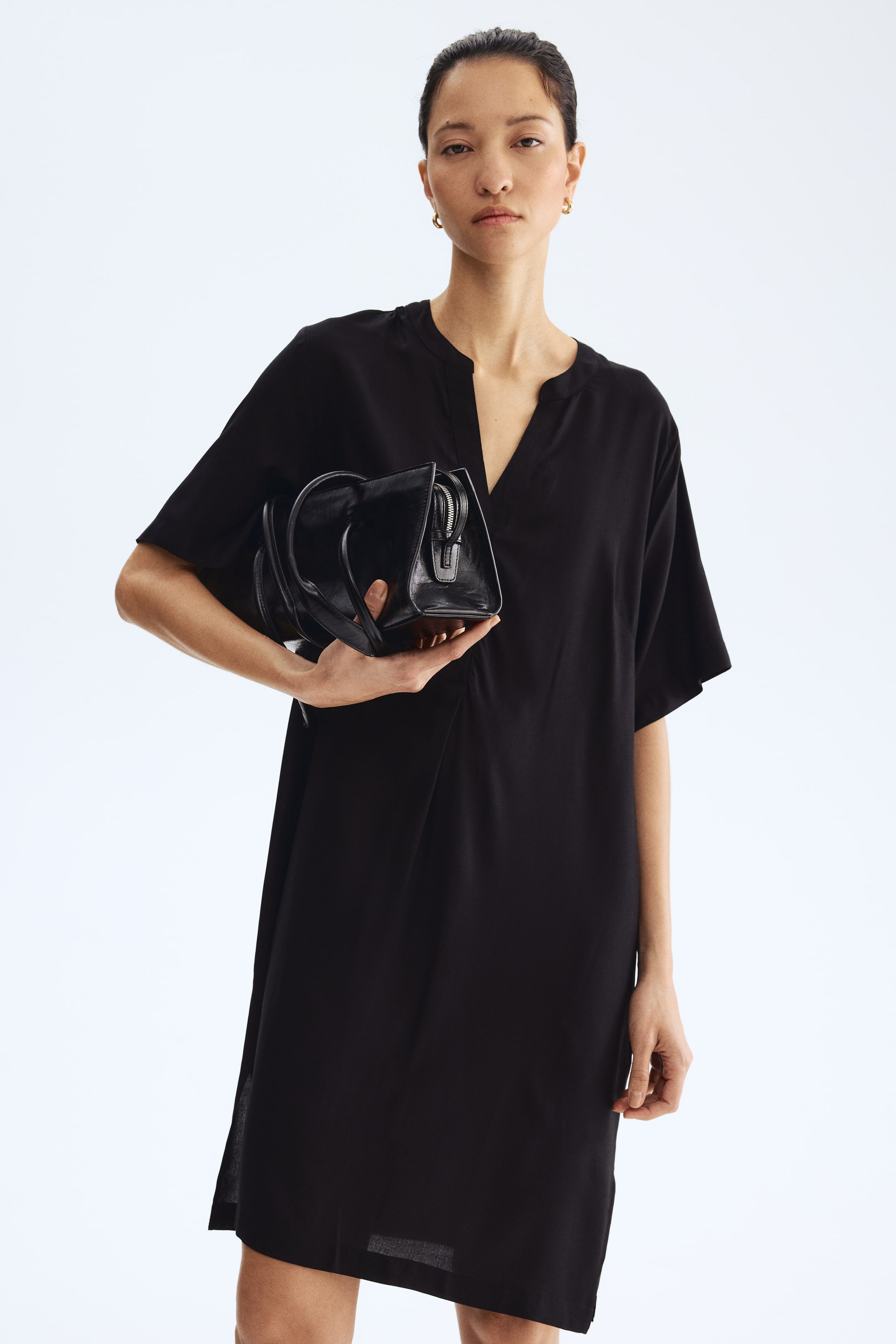 查看大圖: 這款黑色連衣裙採用寬鬆直筒剪裁，無領V字領口，短寬袖，正面縫線細節。裙擺落於膝上，搭配一個黑色手提包。