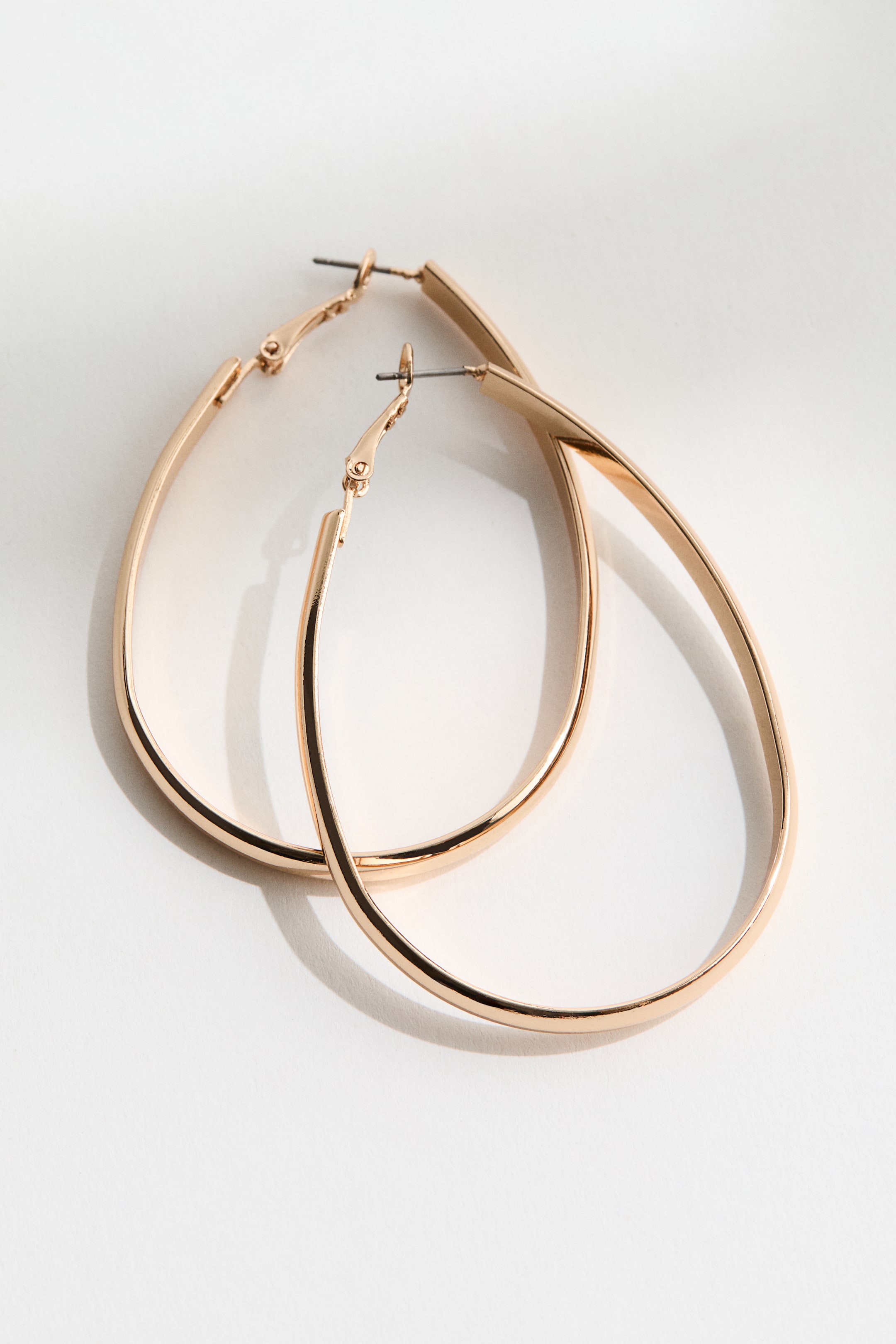 Agrandir l'image: Deux boucles d'oreilles créoles dorées reposent, l'une chevauchant partiellement l'autre. Elles présentent une forme ovale allongée avec des côtés aplatis et des fermoirs à tige visibles.