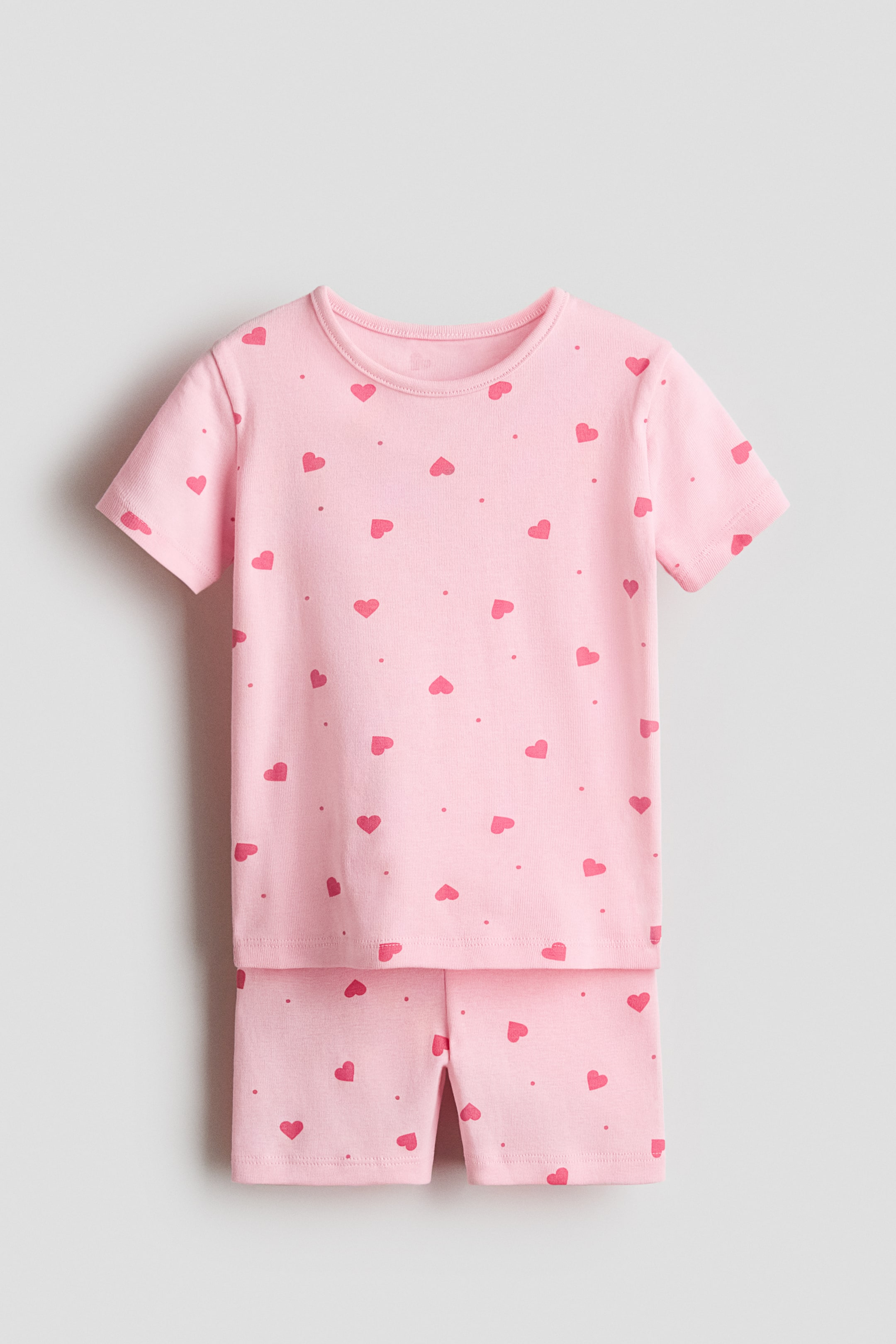View larger image: A pink pajama set is laid flat, featuring a T-shirt and matching shorts, both displaying an all-over print of darker pink hearts and small dots. The T-shirt has a round, narrow-trim neckline and short sleeves, while the shorts have an elastic waistband.