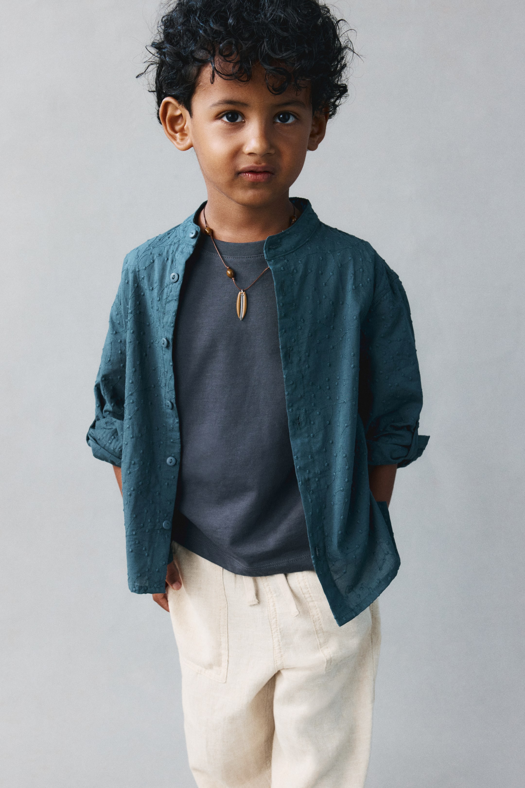 View larger image: Cream-colored loose-fit pants in an airy cotton weave are shown from the front, featuring a relaxed drape with tapered legs. The pants have an elasticated waist with a visible cream drawstring and patch front pockets, styled with an overlapping dark grey t-shirt and an open teal button-up shirt.