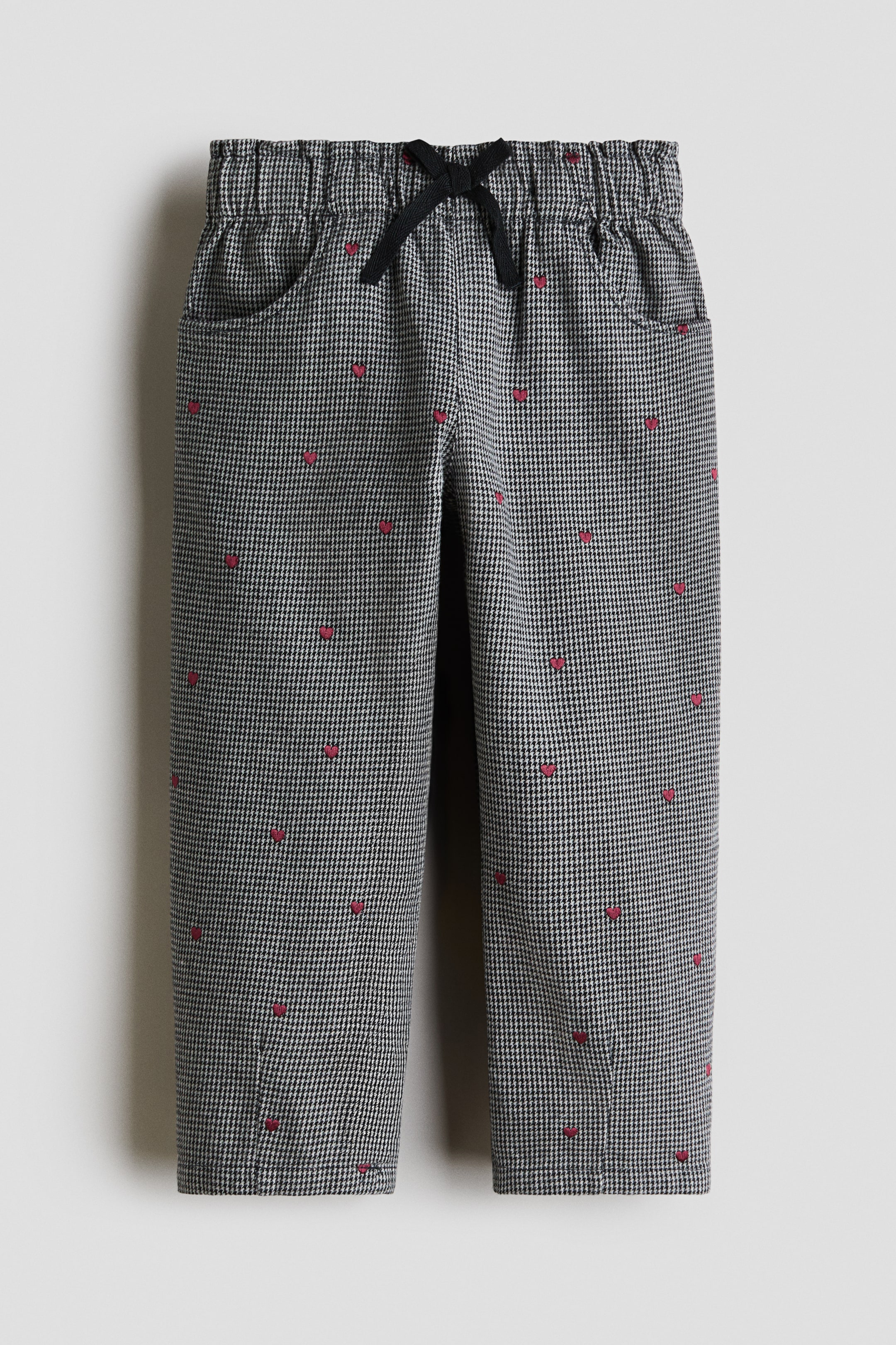 Grotere afbeelding bekijken: Een broek, van voren weergegeven, met een zwart-wit pied-de-poulemotief versierd met kleine, verspreide rood geborduurde hartjes. De broek heeft een elastische tailleband met een zwart trekkoord in strikvorm en steekzakken aan de zijkant.