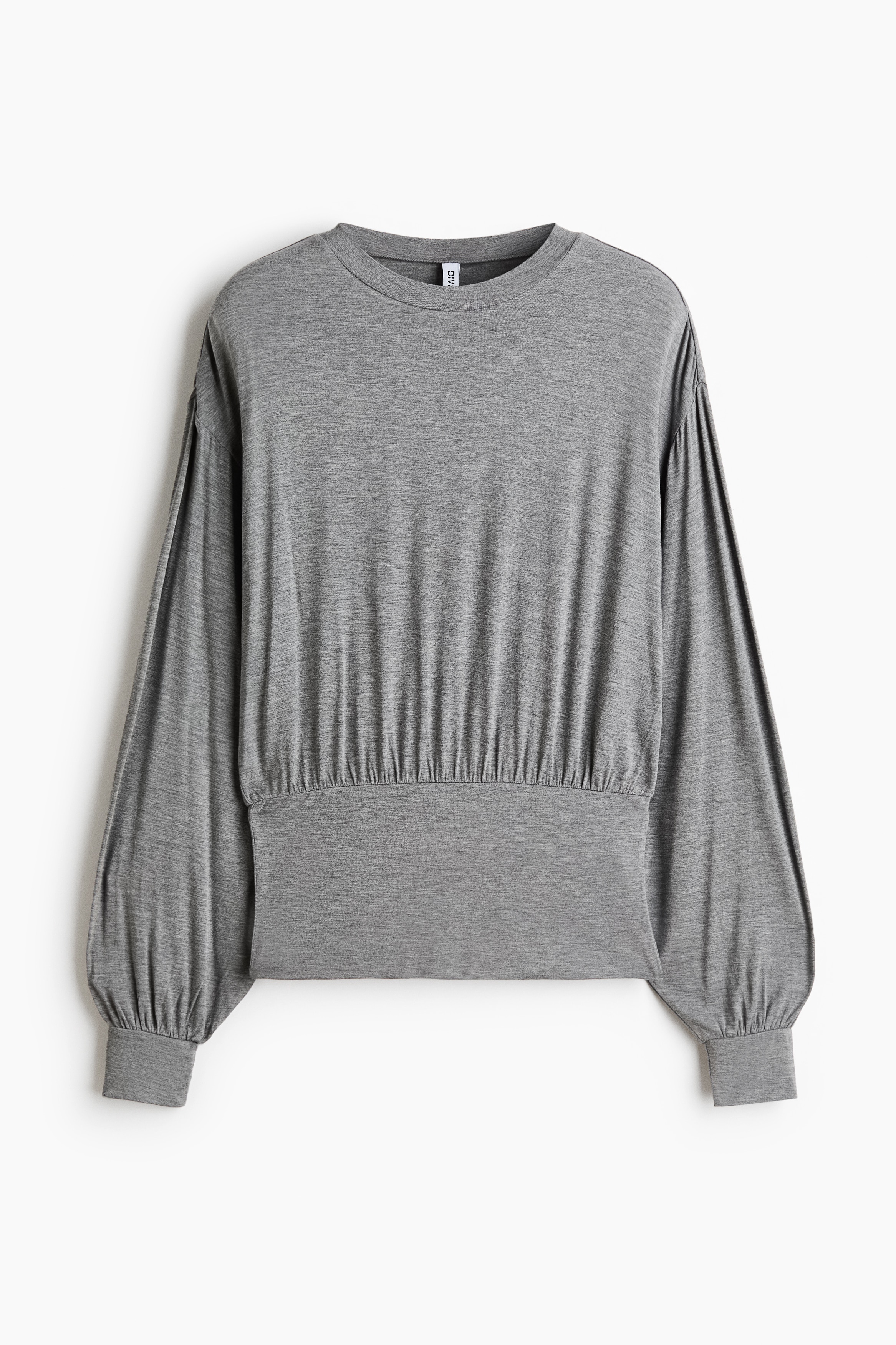 La modelo lleva una prenda superior gris de manga larga con cintura ajustada y fruncida, y mangas de corte relajado, de pie de perfil con los brazos levantados. La prenda se combina con unos vaqueros azules de tiro bajo y lavado claro.