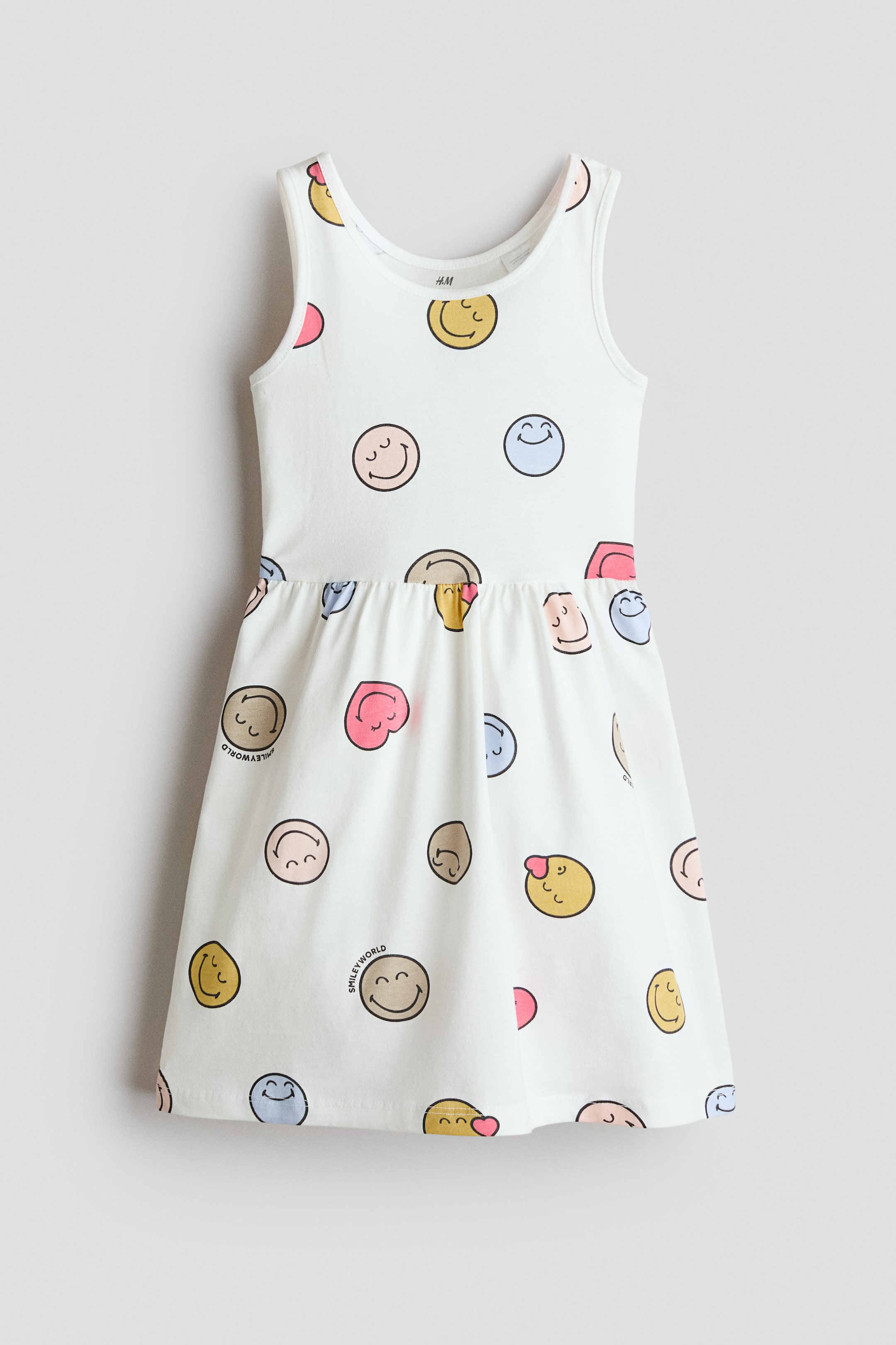 View larger image: The white sleeveless dress features a colorful pattern of assorted smiley faces, some with "SMILEYWORLD" text. It is designed with a round neckline, a fitted bodice, and a gathered waist that flows into a flared skirt.