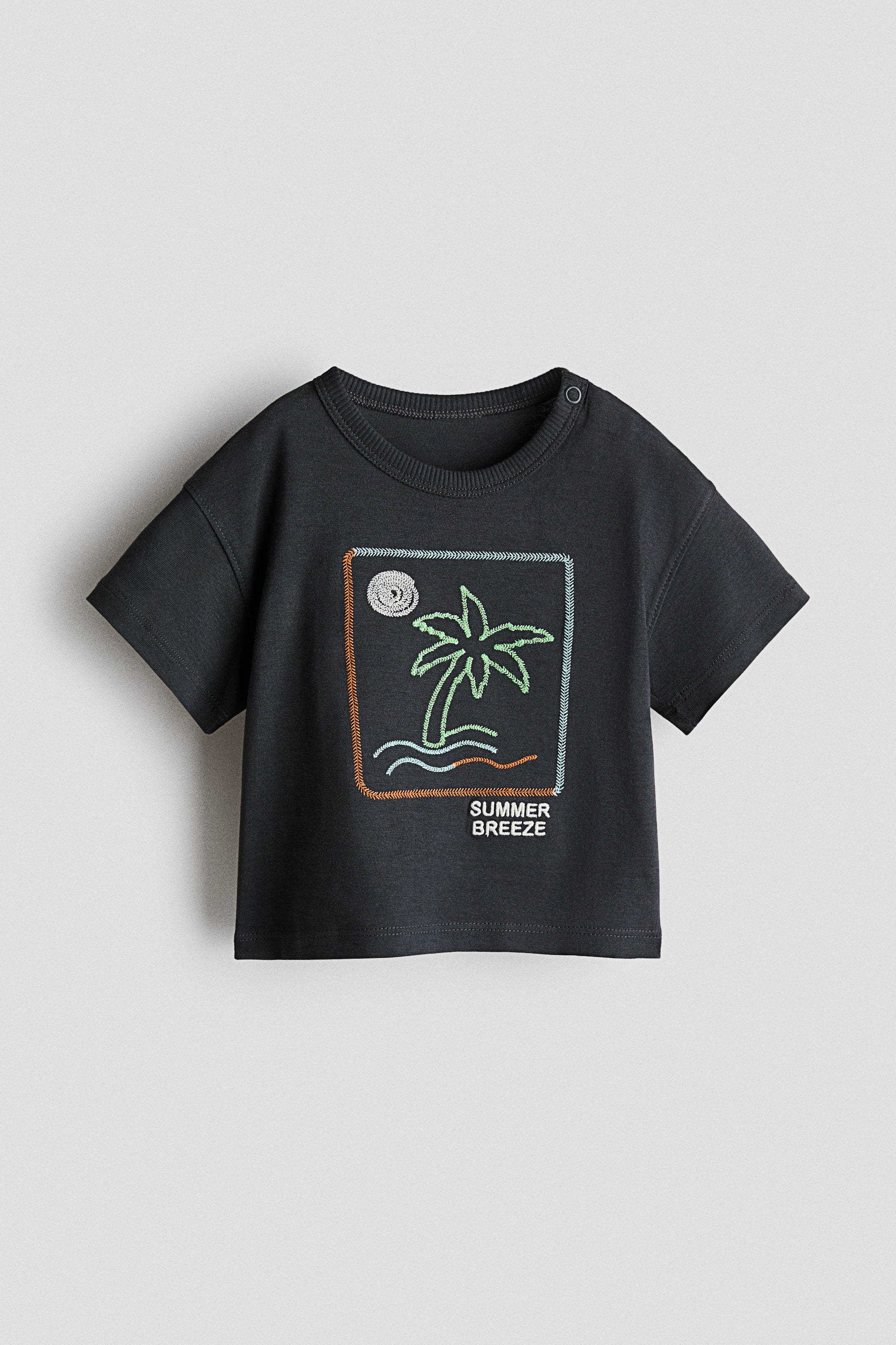 View larger image: The dark grey t-shirt is displayed front-facing, featuring short sleeves and a ribbed crew neck with two snap buttons on the left shoulder. A vibrant embroidered design adorns the chest, showcasing a light green palm tree with light blue waves and a white sun, all encased within a dashed rectangular border with the text "SUMMER BREEZE" appearing below.