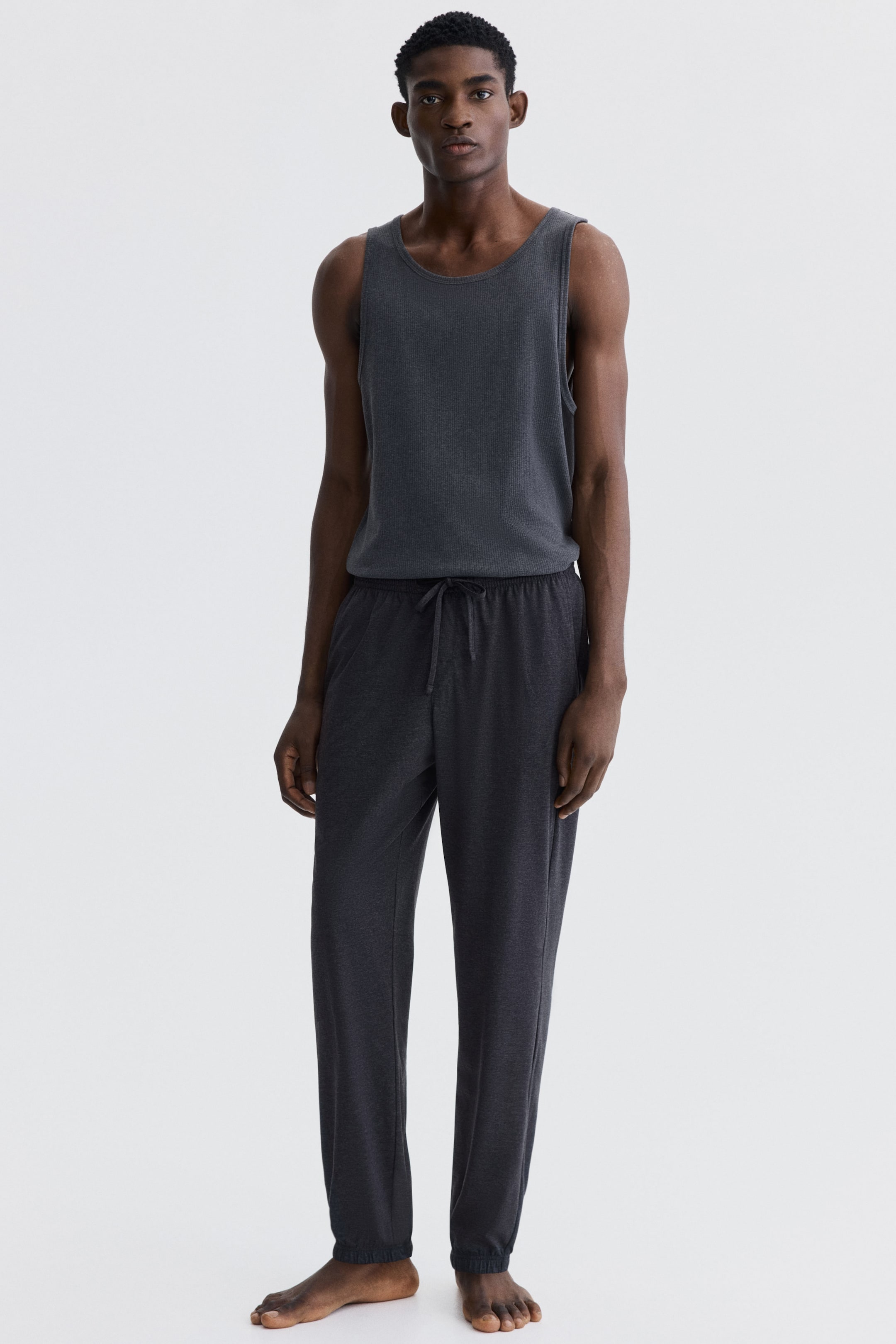 Prikaži veću sliku: Predstavlja se tamno sivi rebrasti pleteni top bez rukava i odgovarajuće tamno sive trenerke. Top bez rukava ima okrugli izrez, a trenerke su dizajnirane sa strukom na vezivanje i elastičnim manžetnama na zglobovima.