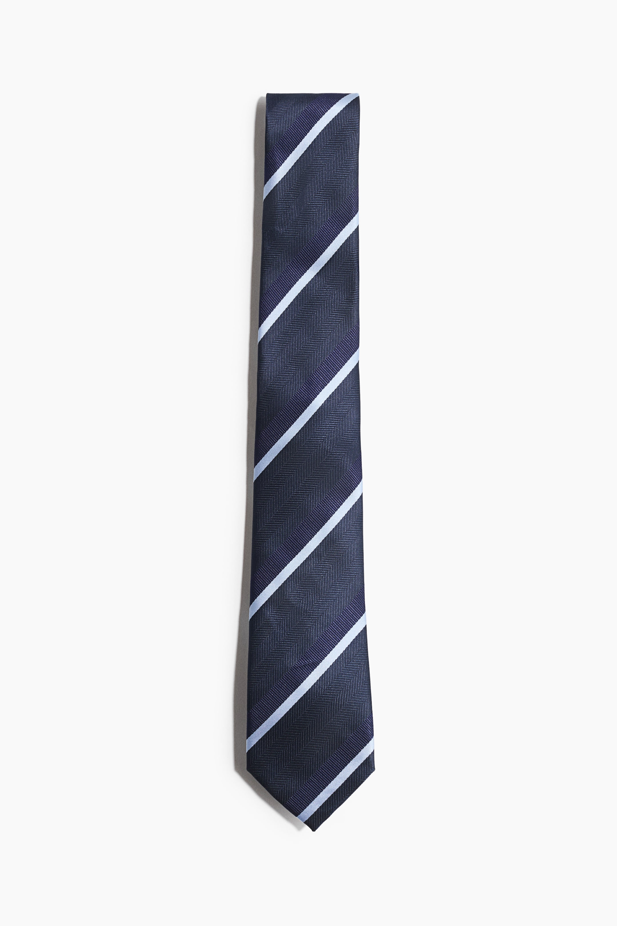 View larger image: This classic tie showcases a dark blue herringbone woven fabric on its front, adorned with diagonal light blue stripes across its surface.