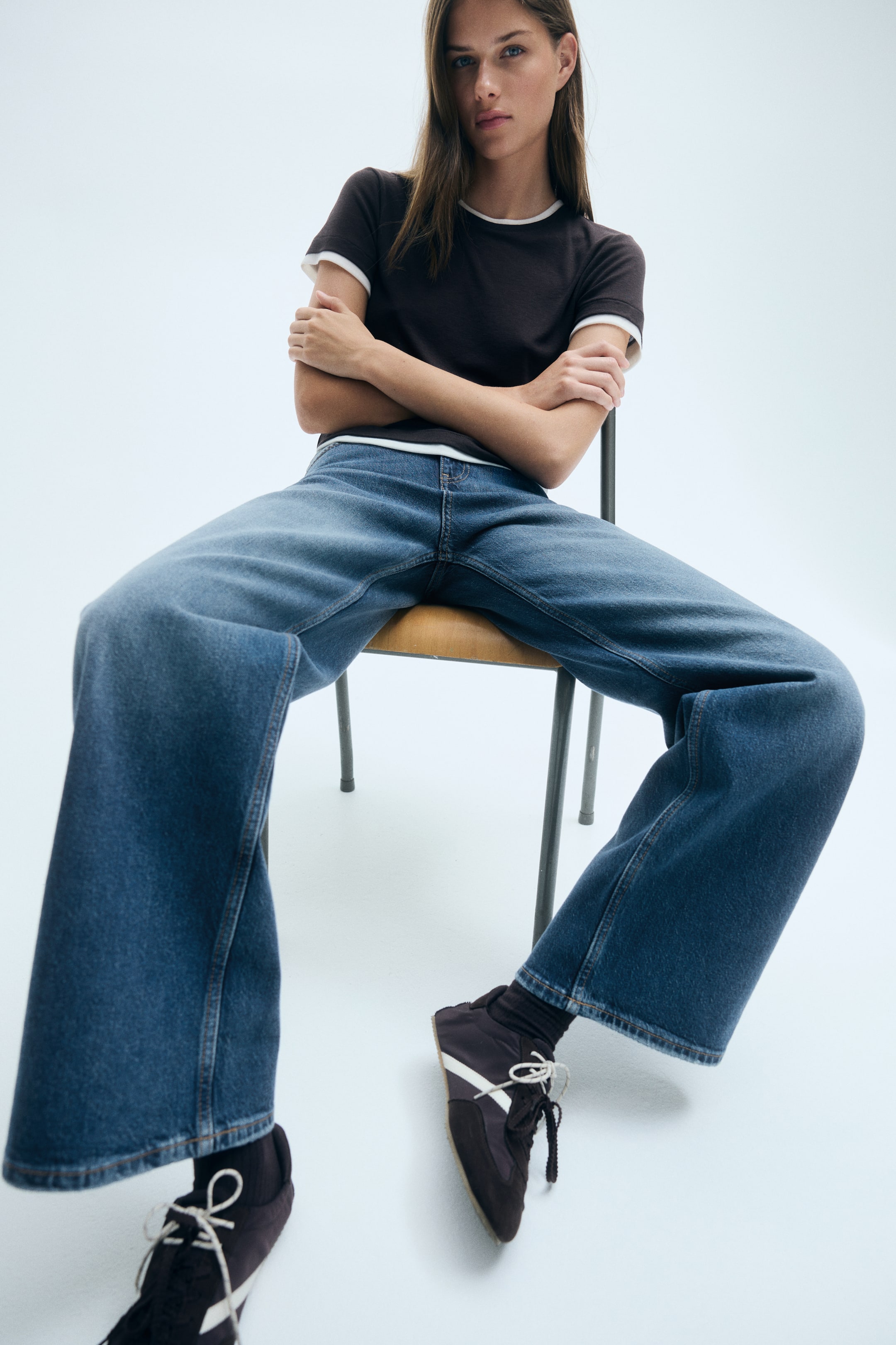 拡大画像を表示: ブルーデニムのジーンズは、ゆったりとしたストレートレッグのシルエットで、椅子に座るモデルが着用しています。ミディアムウォッシュのジーンズは、ダークカラーの半袖Tシャツとロートップスニーカーに合わせてスタイリングされています。
