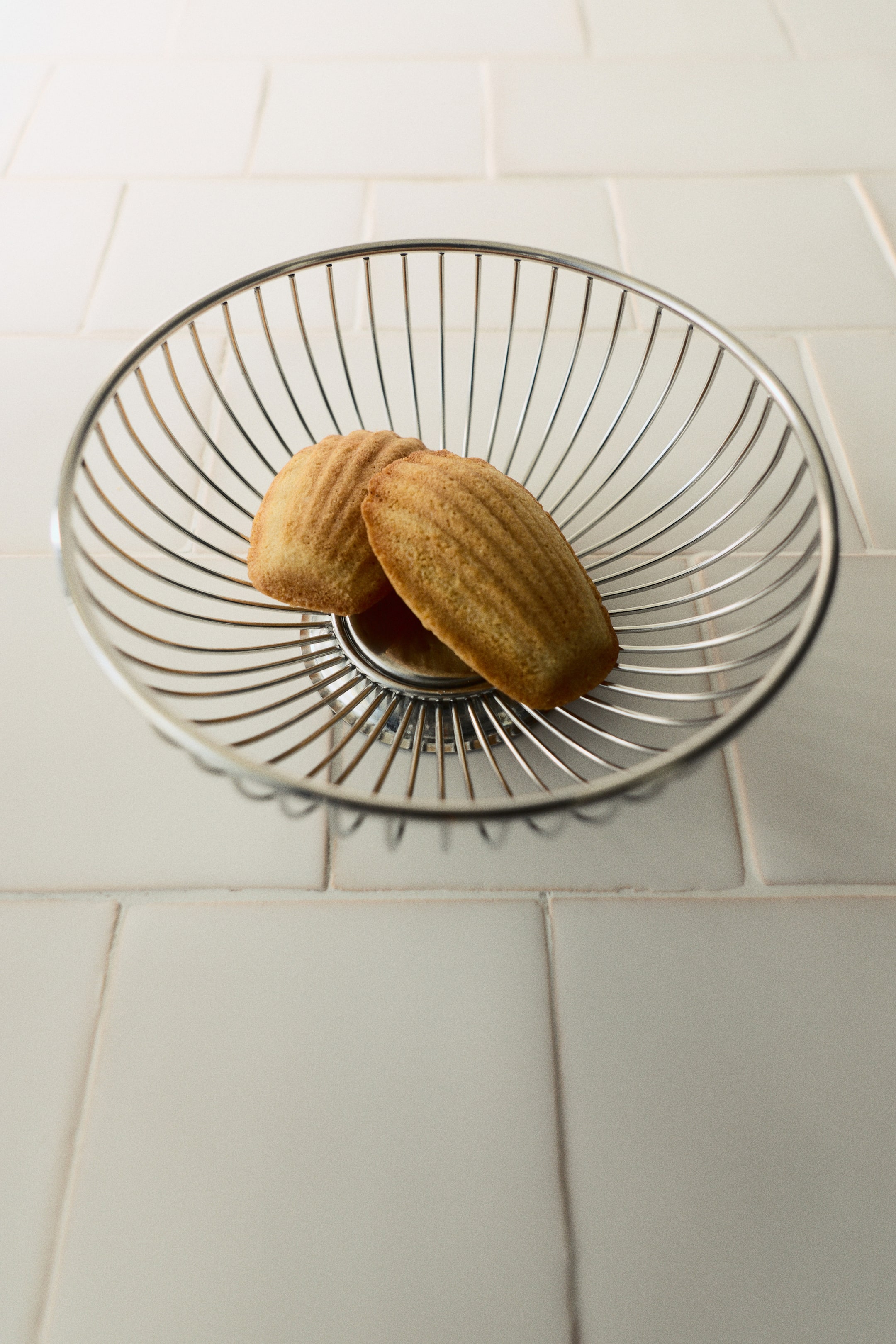 Agrandir l'image: Un petit panier à pain, fabriqué en fil d’acier inoxydable, présente un design minimaliste. Sa structure circulaire est composée de fils rayonnant depuis une base centrale pleine et se courbant vers le haut, tenant deux Madeleines.