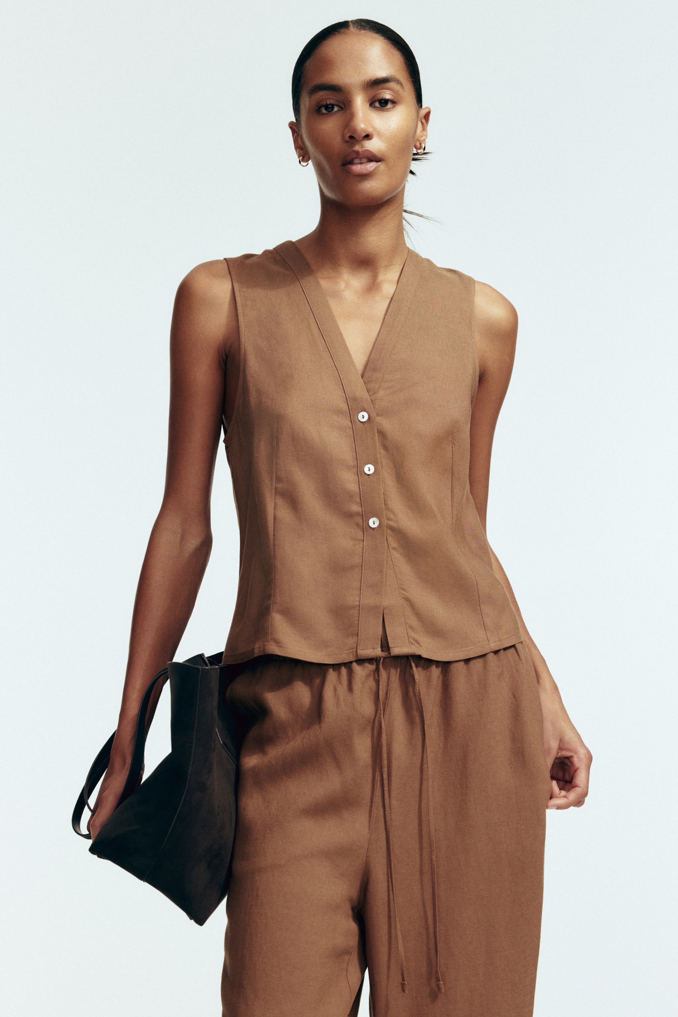View larger image: A brown sleeveless top with a V-neckline and three white buttons down the front is worn by the model. The top features front shaping seams and is styled with matching brown pants.
