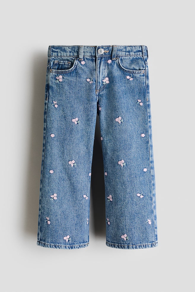 Blue denim jeans are lying flat, presenting a frontal view, featuring scattered pink floral embroidery motifs across both legs. They are designed with two front pockets, a small coin pocket, a waistband with belt loops and a silver-toned button closure, and display a loose fit with wide legs. - 1