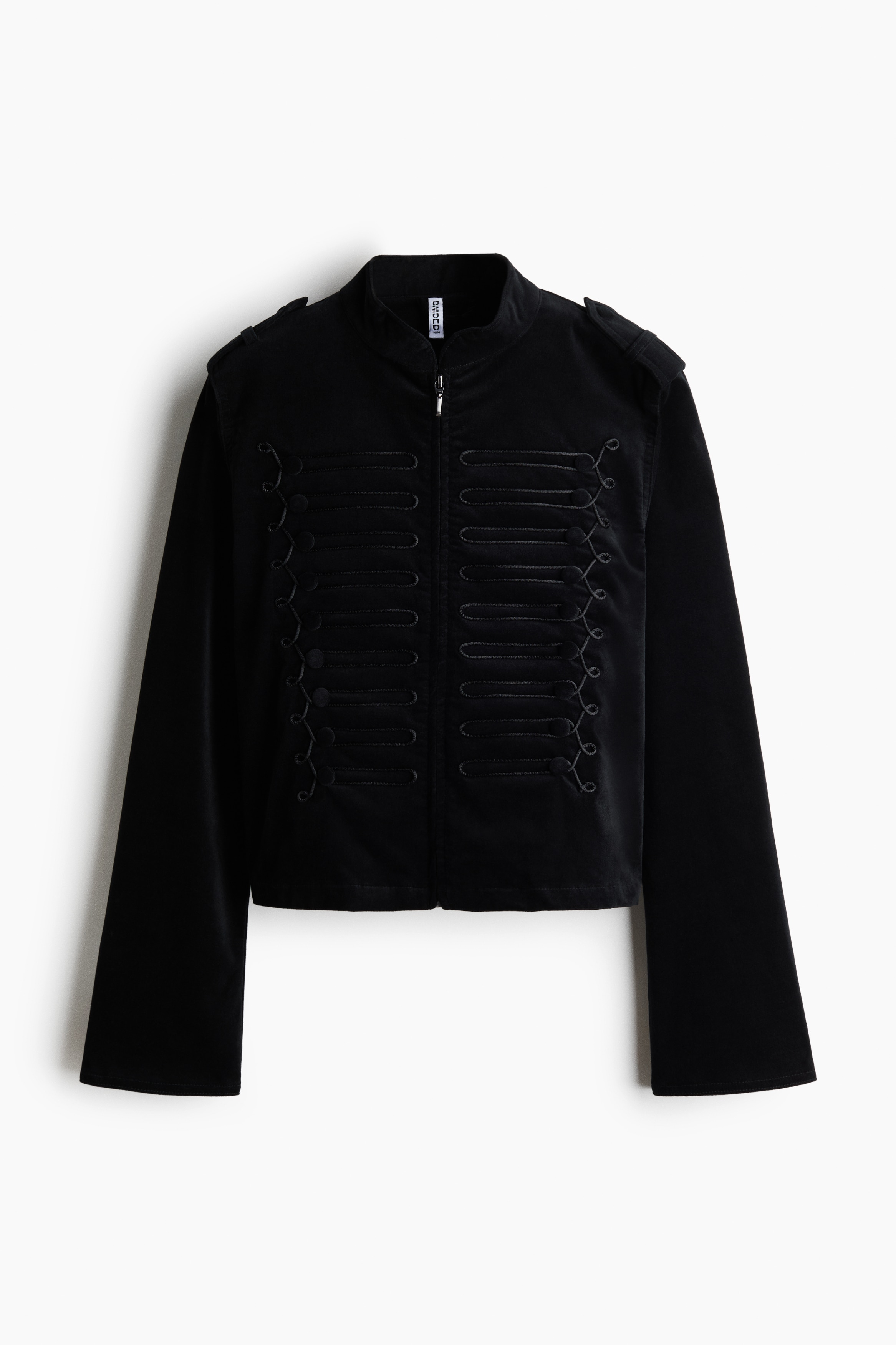 The model is wearing a black cotton velvet jacket featuring a band collar, a partially open front zip, and decorative horizontal military-style loops with covered buttons on both sides of the chest. The short, fitted jacket is styled with a silver chain necklace and paired with light blue jeans.