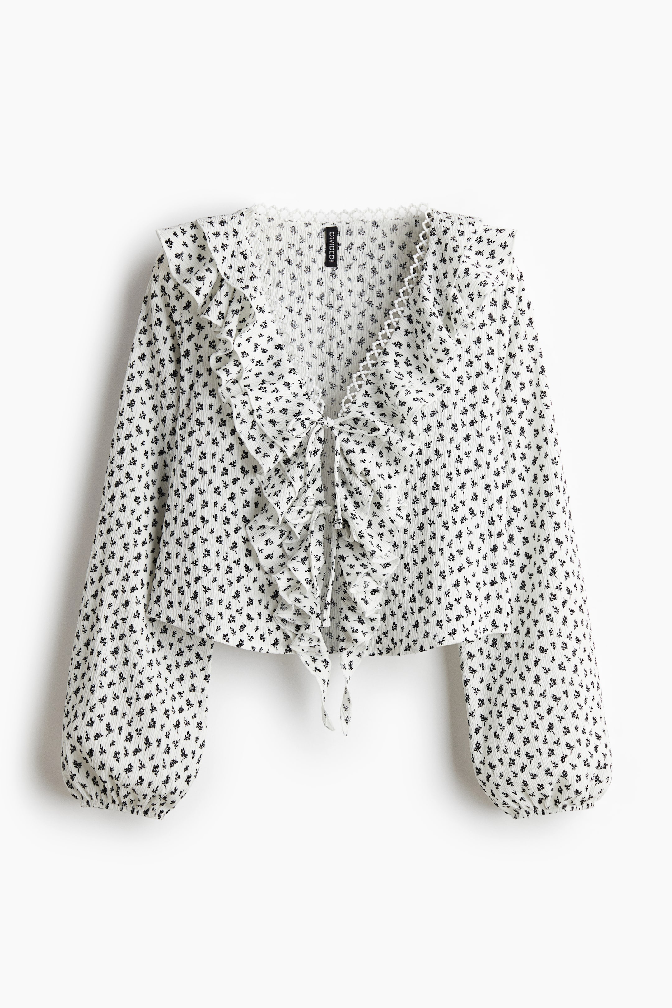View larger image: A white blouse with a small black floral print and a subtle crinkled texture is presented FRONT. It is featuring a ruffled V-neckline with a scalloped edge and a tie-front closure, and long, voluminous sleeves that are gathering at the cuffs.