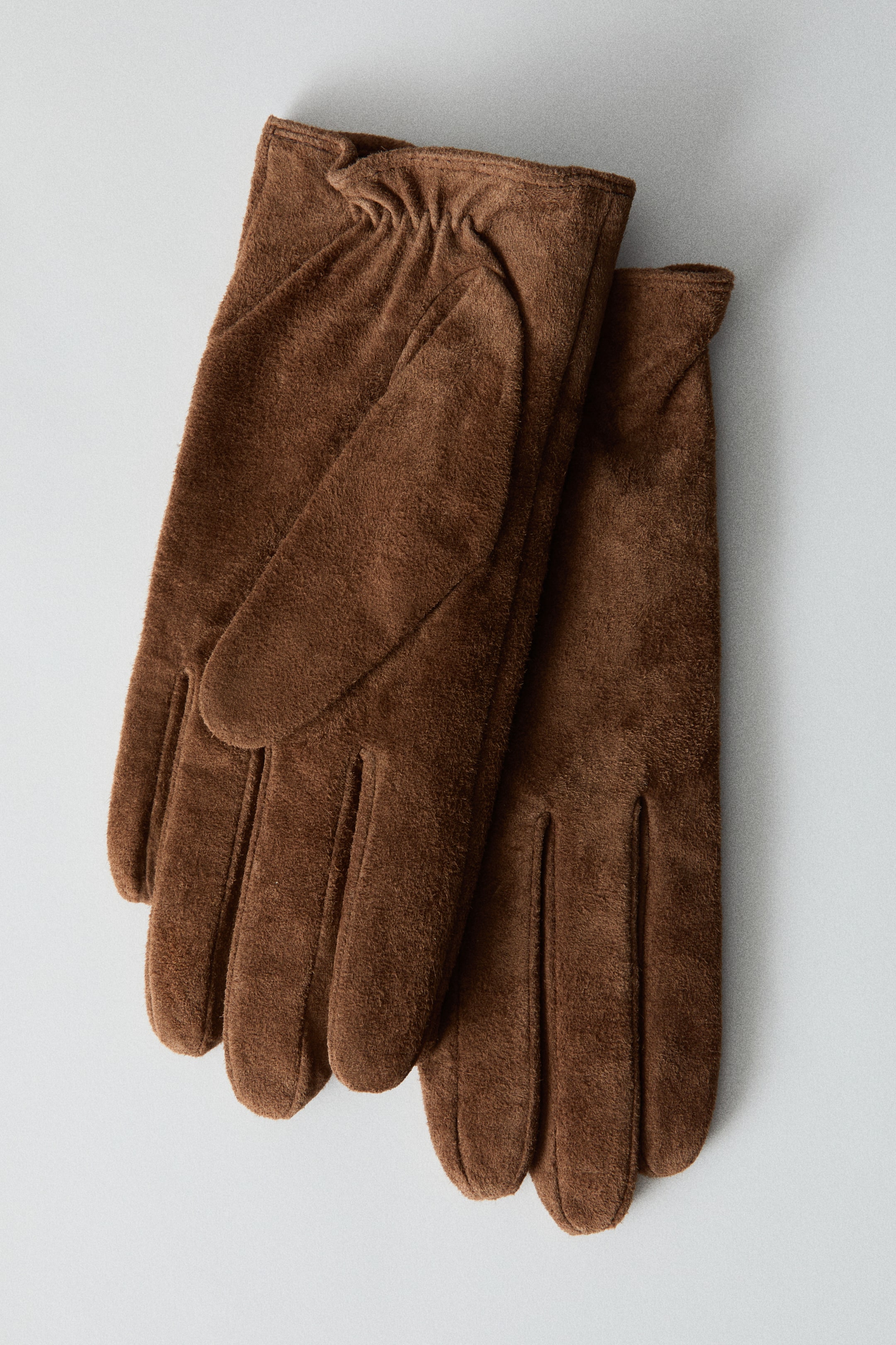 Ver imagen más grande: Un par de guantes de ante marrón está colocado en posición horizontal, con uno superpuesto al otro para mostrar ambas caras. El guante superior presenta un detalle fruncido elástico en la muñeca, mientras que ambos guantes muestran costuras limpias y pespunteadas a lo largo de los dedos.