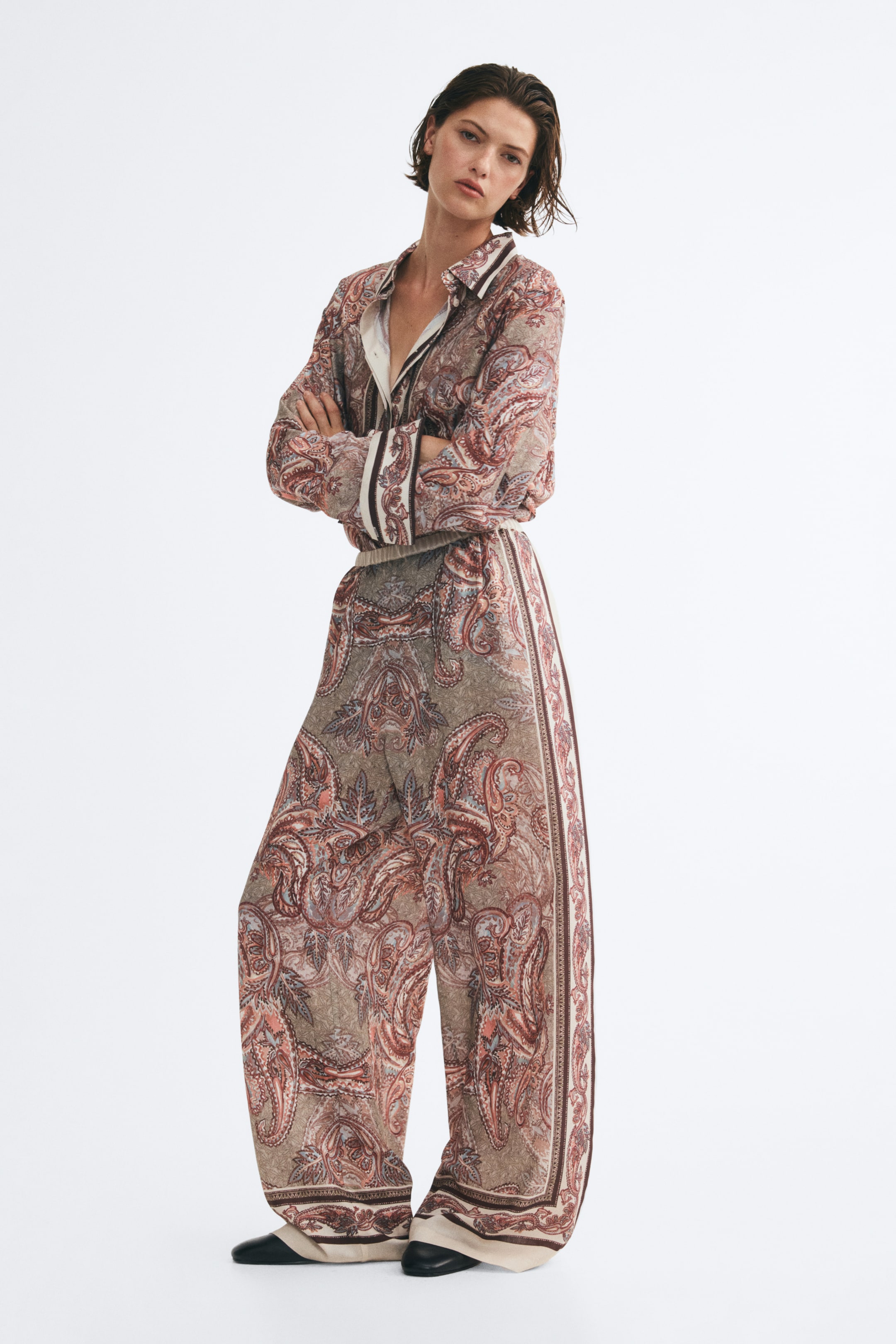View larger image: Loose-fit, wide-leg pants featuring an intricate paisley pattern in brown and beige tones are worn by the model. The high-waisted pants feature a visible elastic waistband and are styled with a matching long-sleeve shirt tucked in.