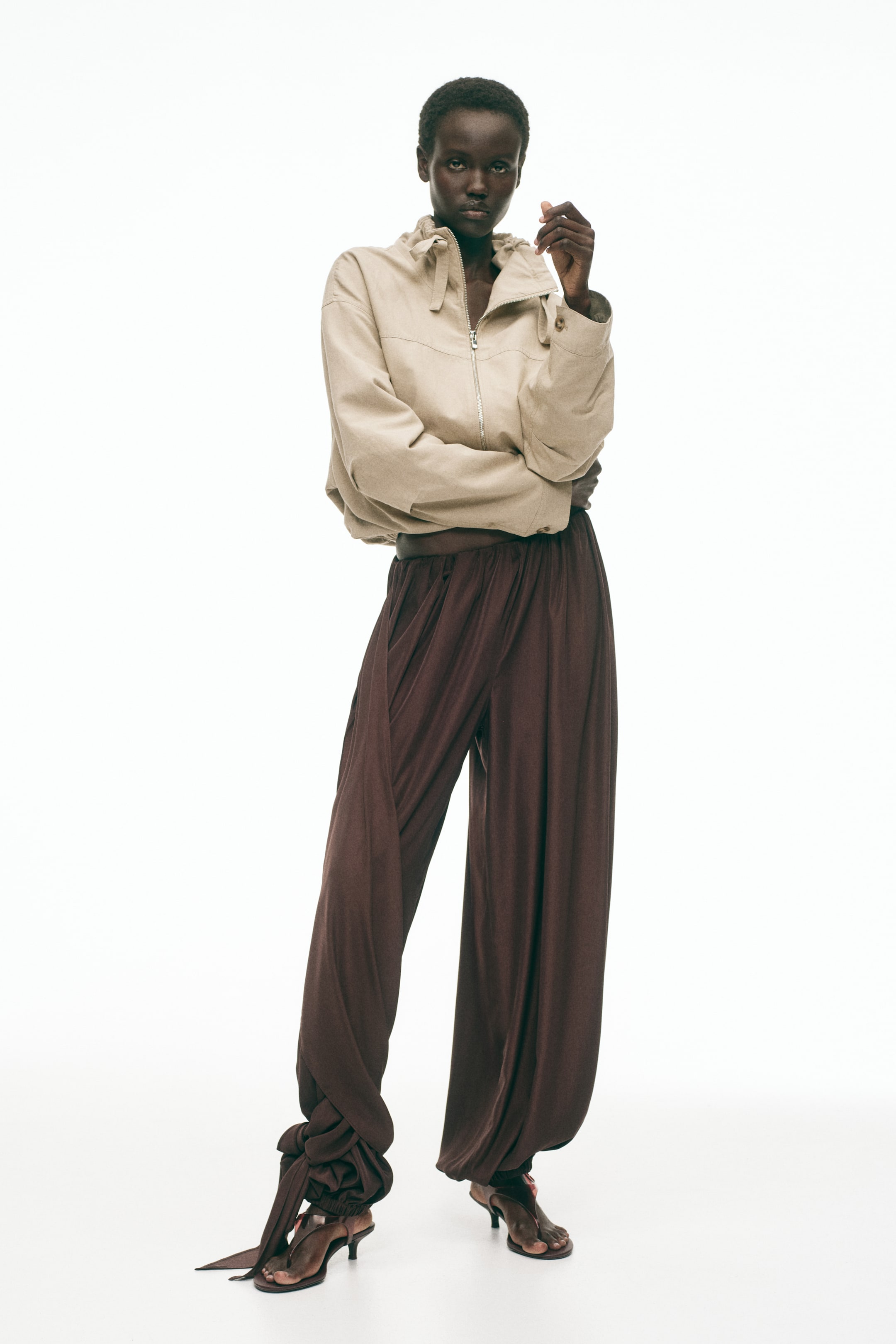 View larger image: A light beige bomber jacket features a stand-up collar with a drawstring, a front zip fastening, and an elasticated hem, whilst dark brown wide-leg trousers showcase a high waist and gathered ankle ties. Dark brown kitten-heel sandals complete the ensemble.