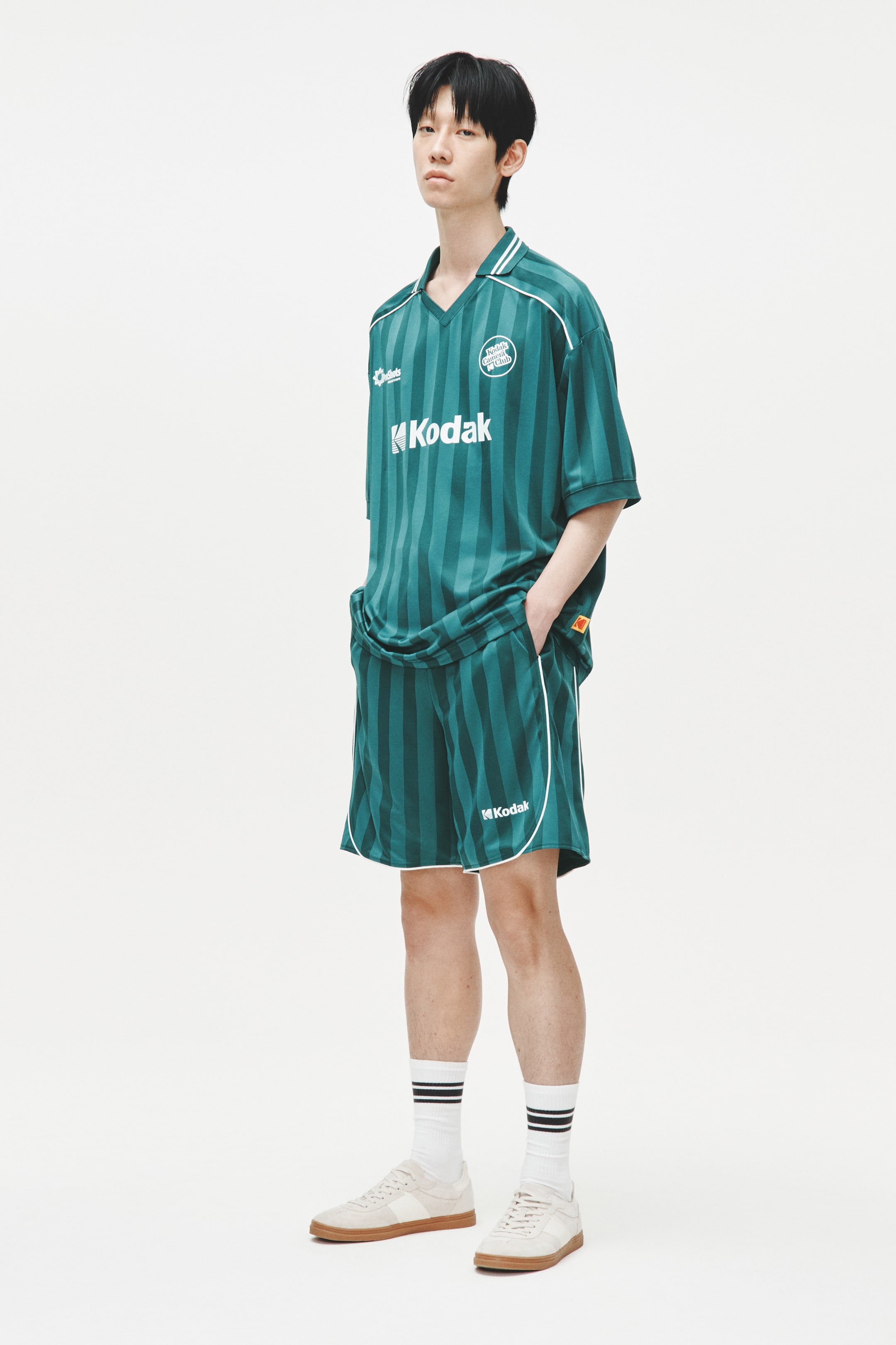 View larger image: A matching teal dan hijau gelap berjalur menegak set jersi atletik dan seluar pendek dipaparkan dari pandangan hadapan yang sedikit menyerong. Jersi ini menampilkan kolar V-neck bertrim putih, lengan pendek dengan trim putih, dan penjenamaan "Kodak" pada dada. Seluar pendek direka dengan paip putih di sepanjang jahitan sisi dan hem, turut menampilkan logo "Kodak" pada kaki kiri.