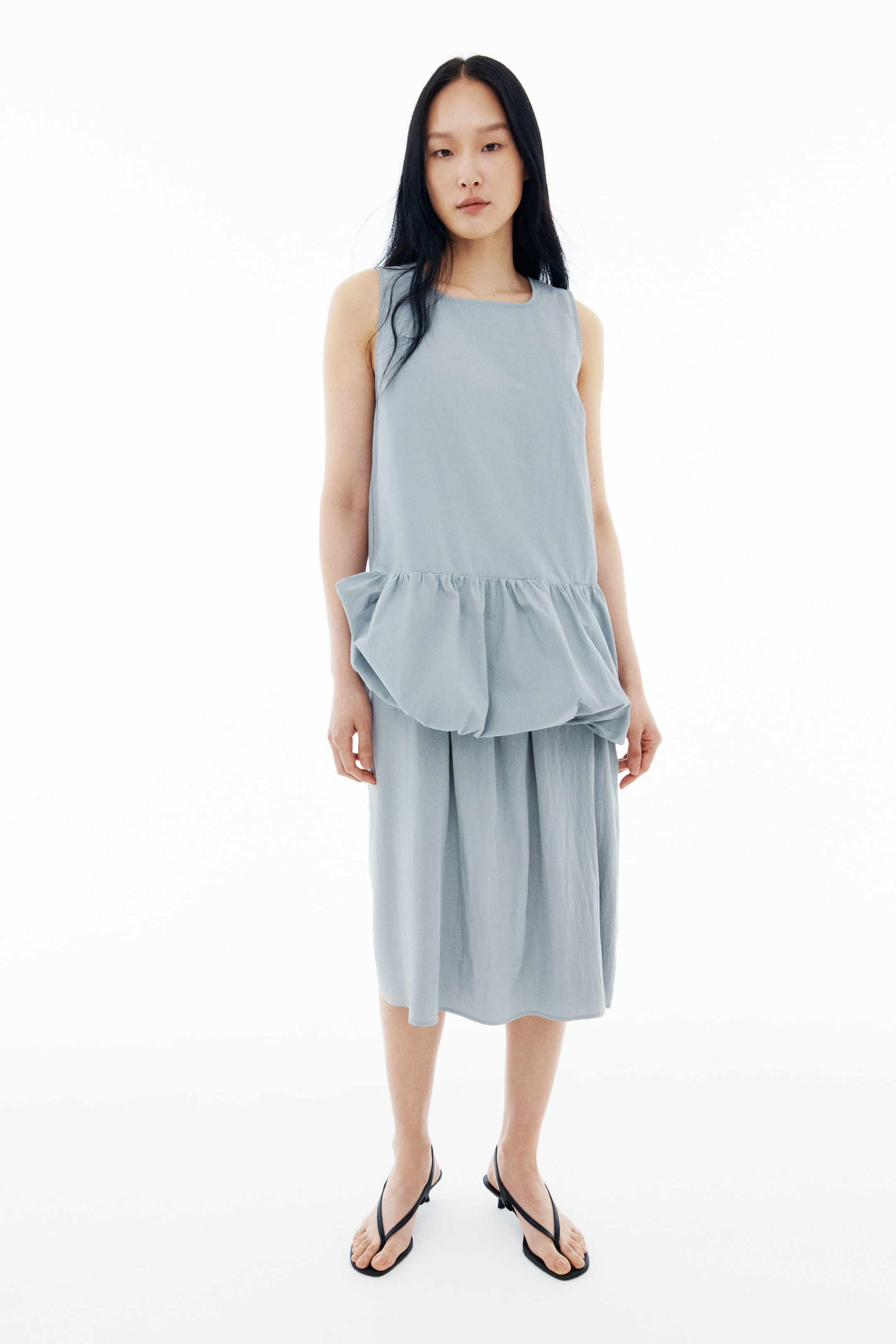 View larger image: The model is wearing a long, light blue sleeveless top with a square neckline. It features a voluminous section at the hem, accentuated by a gathered seam above, forming a bubble hem.