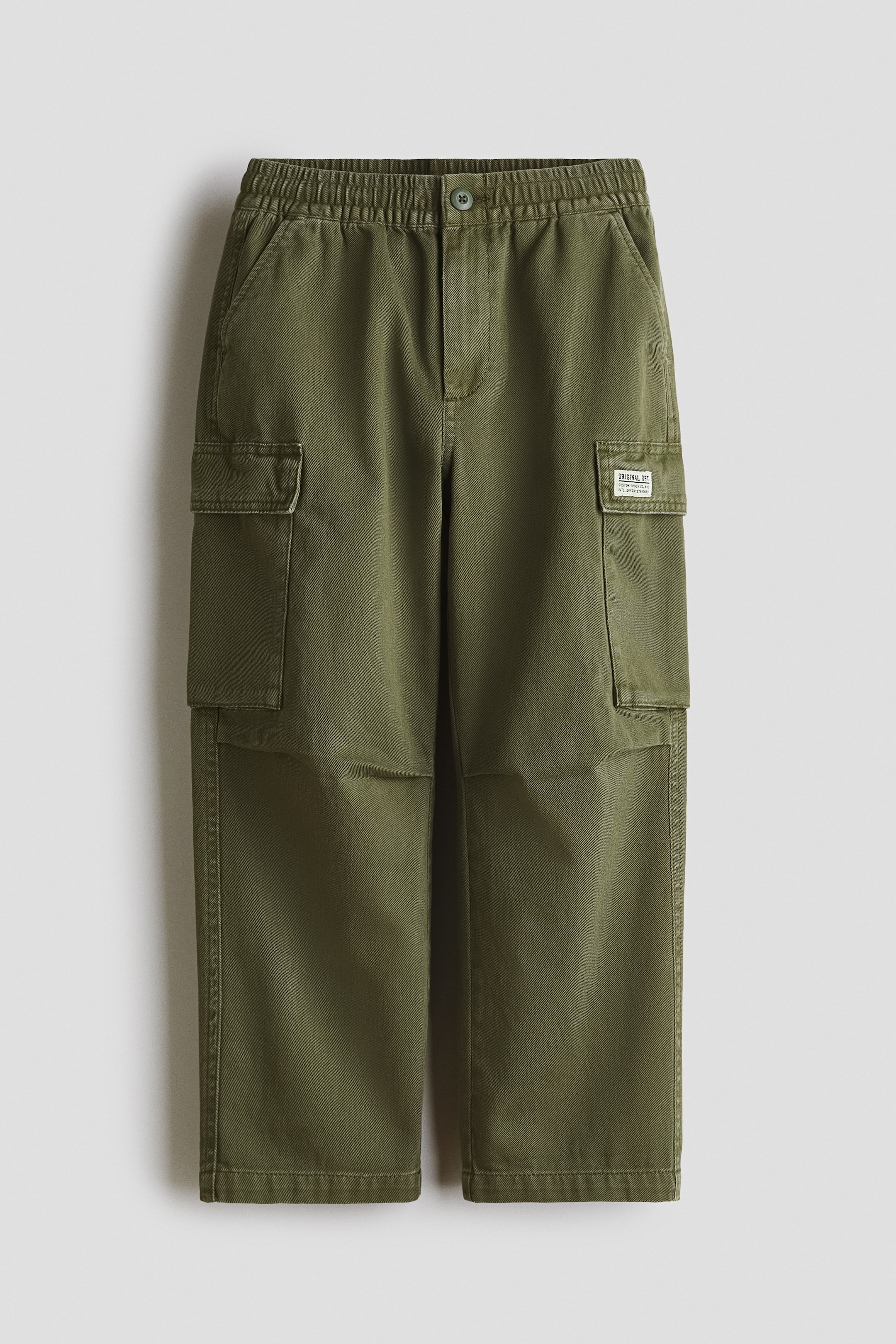 View larger image: Olive green cargo trousers in cotton twill feature an elasticated waist with a button closure and zip fly, along with diagonal front pockets. Patch leg pockets with flaps are also visible, with a small brand label attached to the right leg pocket, all presented from a front flat lay.