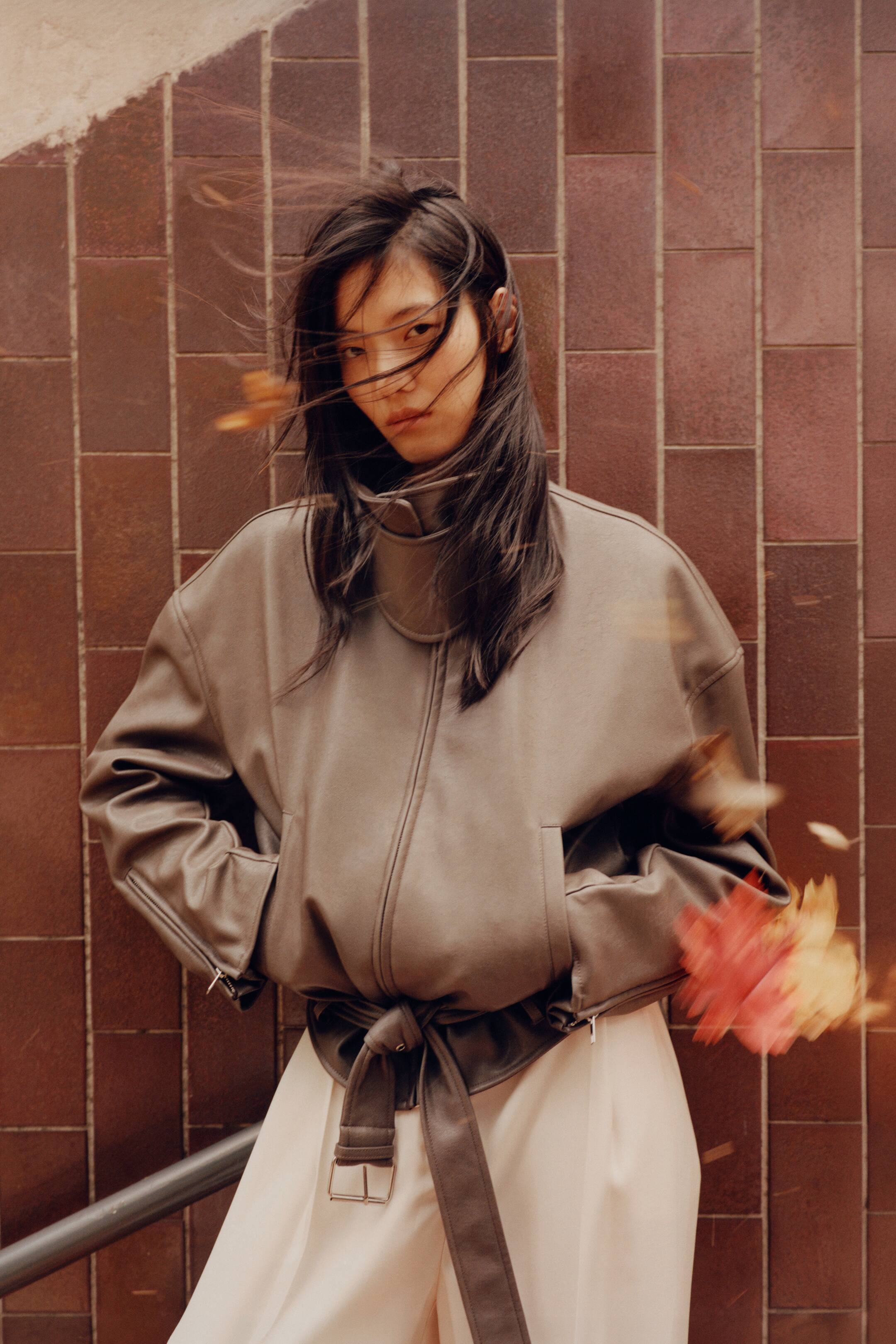 View larger image: An oversized biker jacket in taupe coated fabric is worn by the model, with their hands resting inside the welt front pockets. The jacket features a high funnel collar with a fastened throat latch, dropped shoulders, and an adjustable belt with a metal buckle hanging from the hem, styled over cream-coloured trousers.