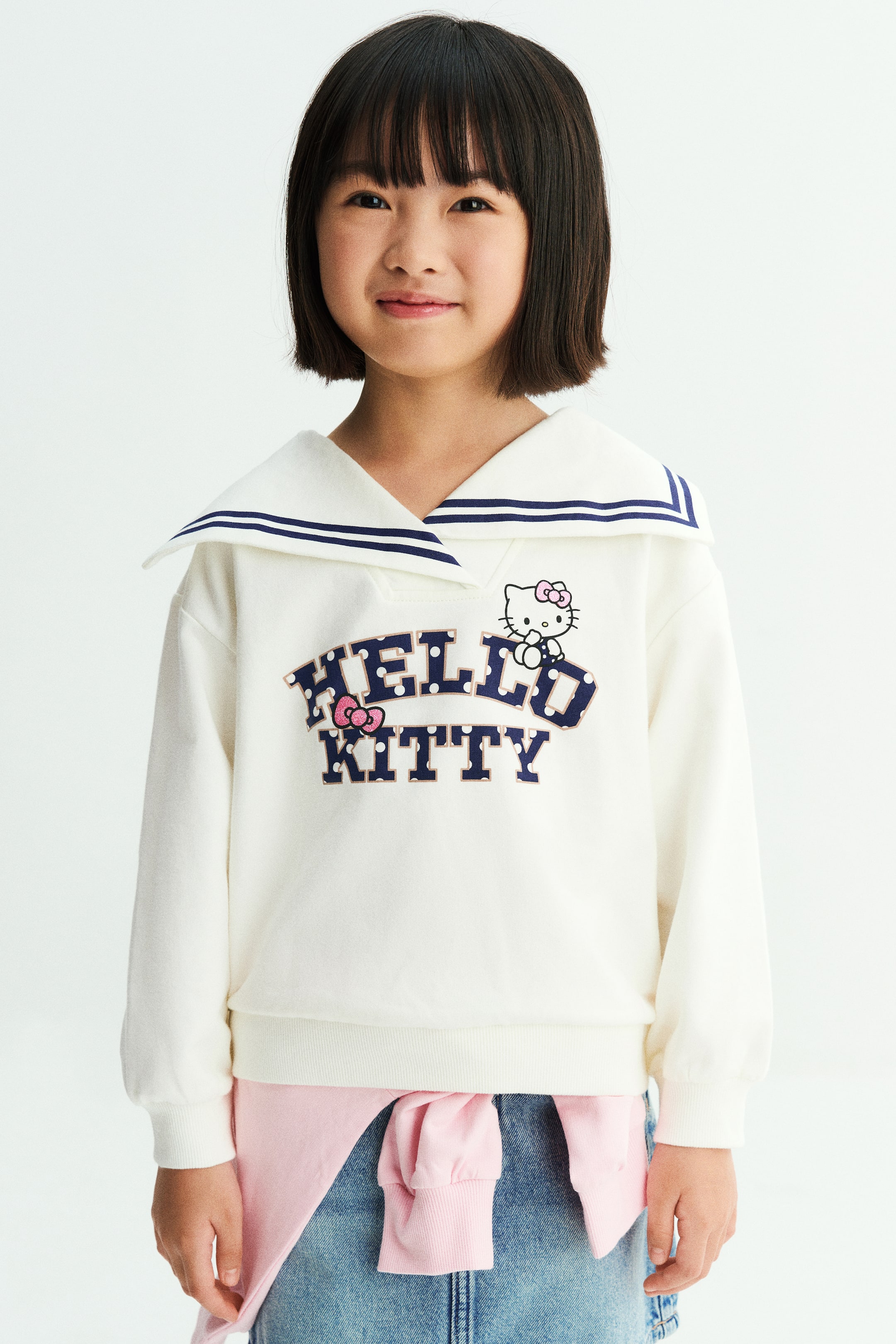 拡大画像を表示: モデルは、ネイビーブルーのストライプが入った大きなセーラーカラーと「Hello Kitty」のプリントグラフィックが特徴のオフホワイトのスウェットシャツを着用しています。Vネックのラップフロント、ドロップショルダー、リブ仕上げのトリムを備え、ライトウォッシュのデニムジーンズと、ウエストに巻いたピンクの長袖シャツを合わせています。