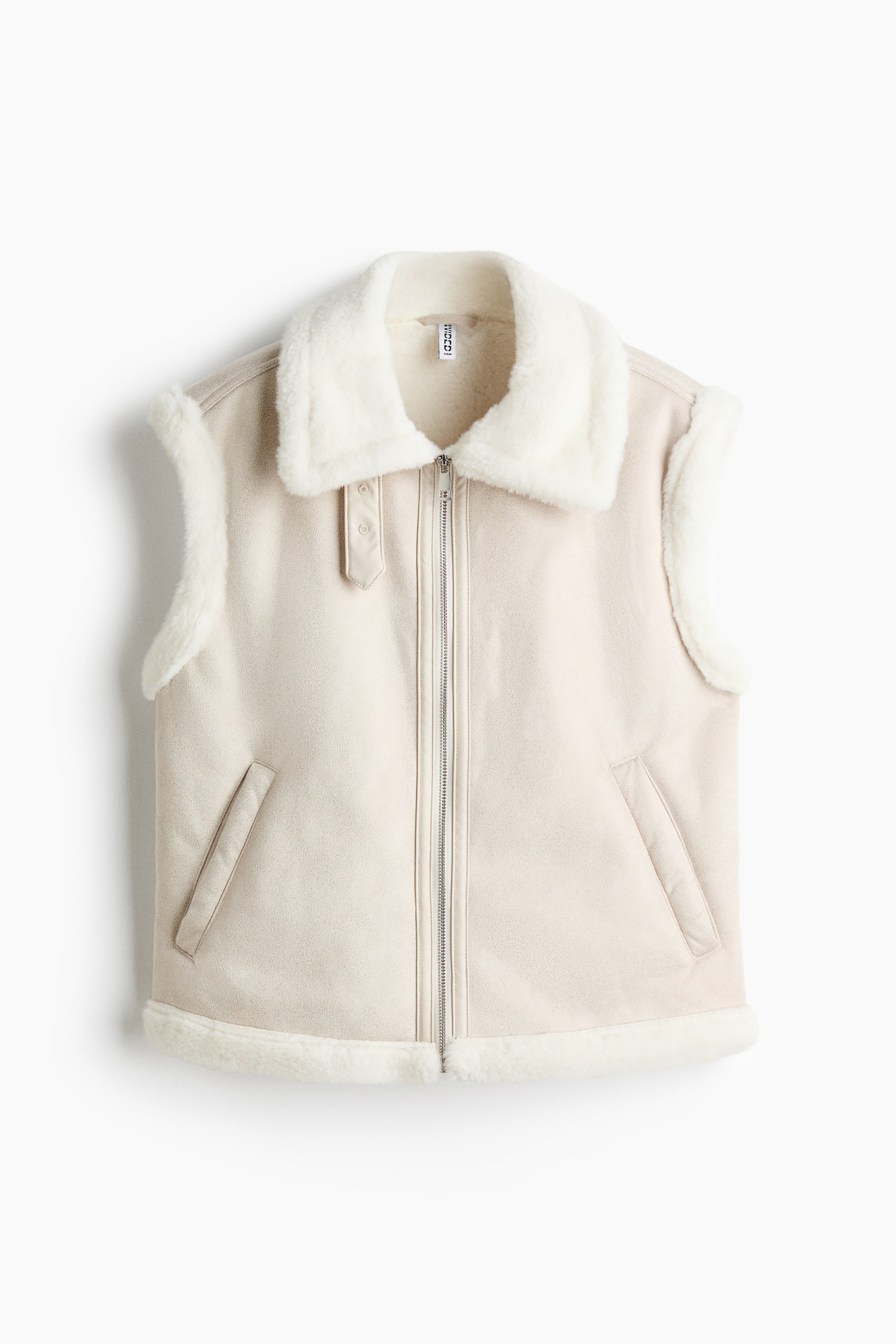 View larger image: A beige suede-look vest is shown from the front, featuring a wide white faux shearling collar and a full-length silver zipper closure. It also shows two diagonal welt pockets, white faux shearling trim along the armholes and bottom hem, and a decorative snap-button strap on the left collar.