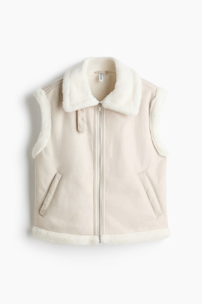 A beige suede-look waistcoat is shown from the front, featuring a wide white faux shearling collar and a full-length silver zip fastening. It also shows two diagonal welt pockets, white faux shearling trim along the armholes and bottom hem, and a decorative snap-button strap on the left collar. - 1