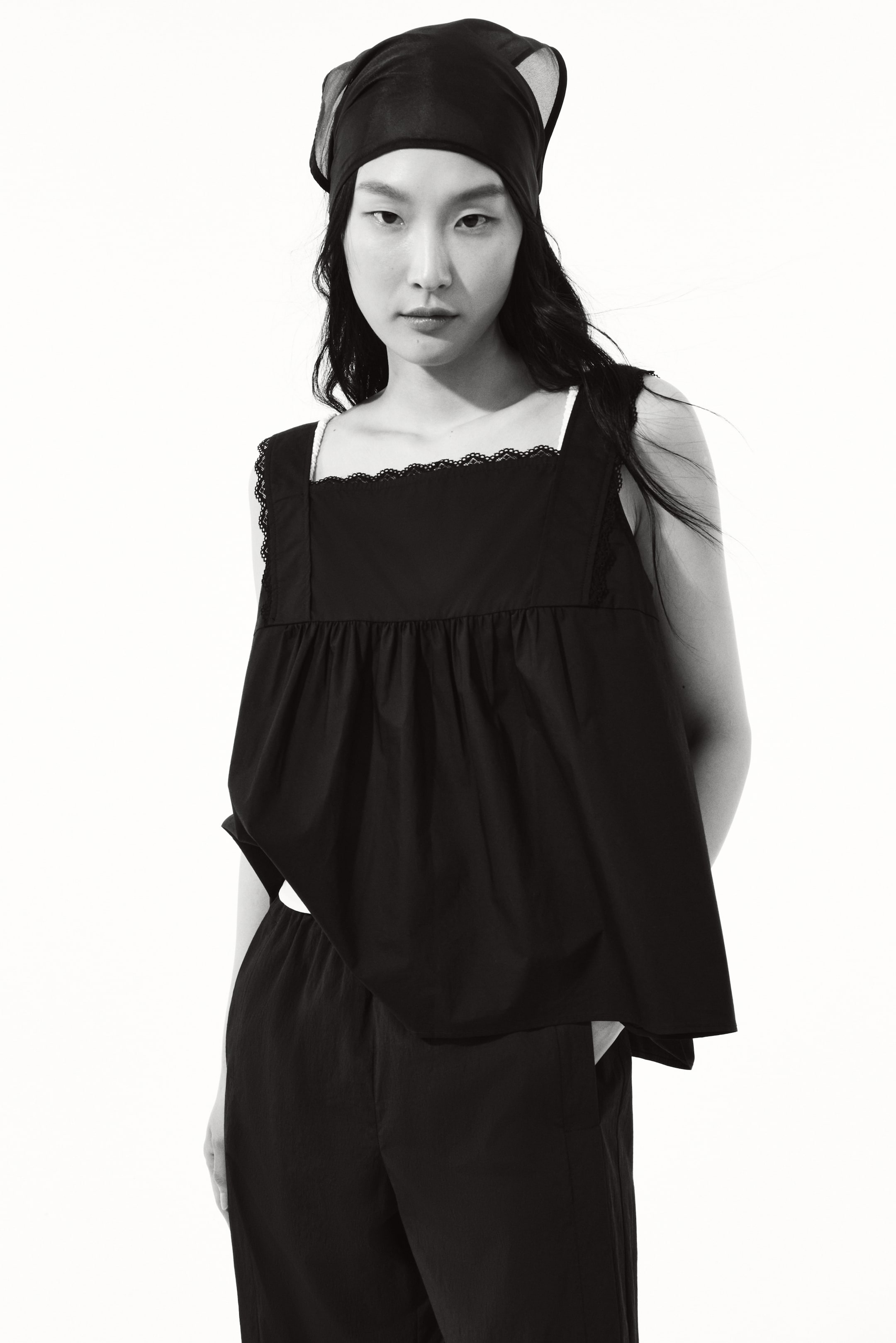 View larger image: Ang modelo ay nakasuot ng itim na pang-itaas na gawa sa magaan na cotton weave. Ito ay mayroong parisukat na neckline na may mga banayad na lace trims, malapad na mga strap sa balikat, at bahagyang A-line na silweta na nilikha ng gathered seam sa harapan.