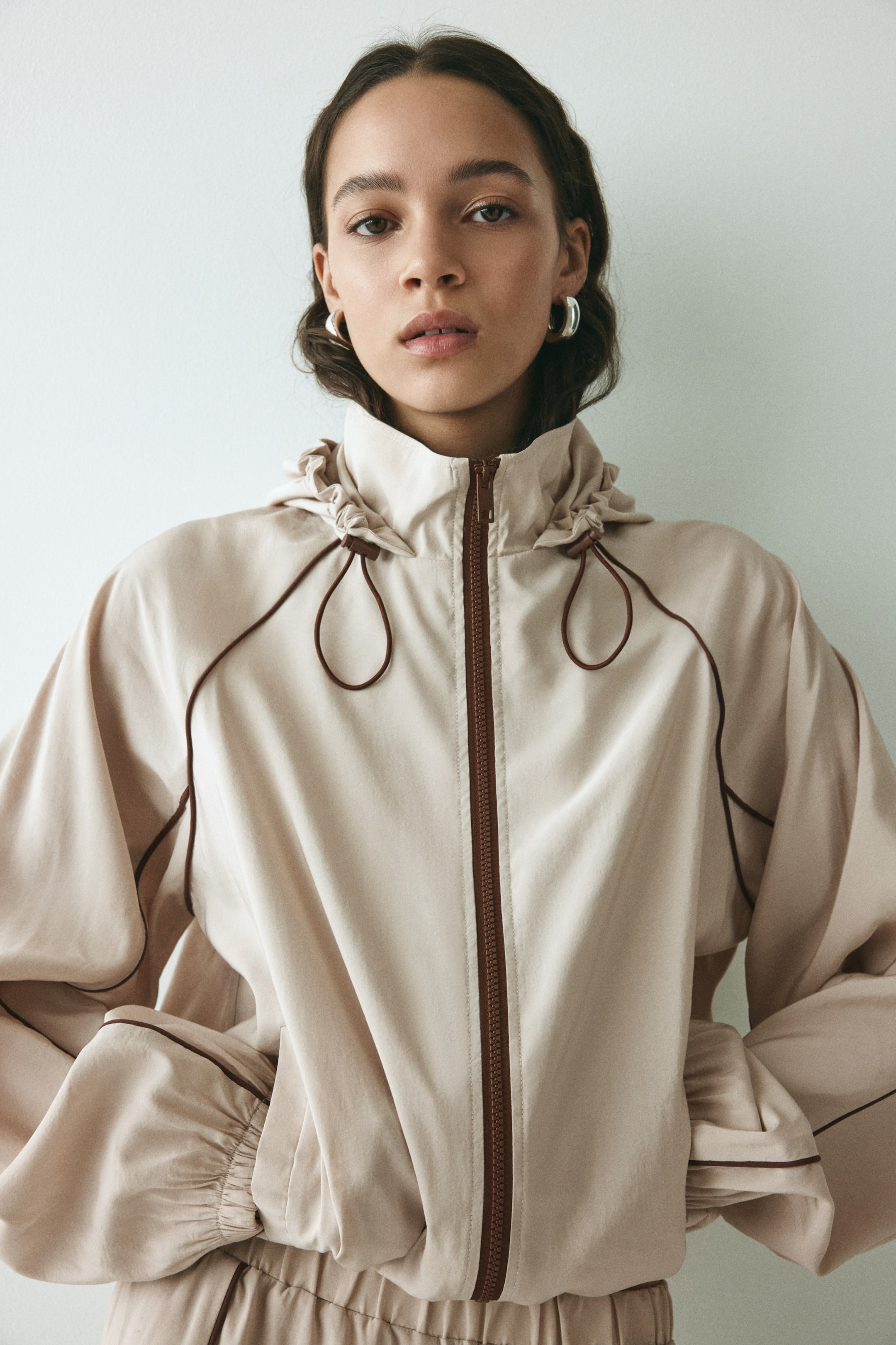 View larger image: A light beige hooded jacket, facing forward, features a full-length dark-brown zipper and dark-brown piping that outlines the raglan sleeves and front seams. A gathered stand collar presents dark-brown drawstrings forming loops, and voluminous sleeves gather at the cuffs, while the bottom hem is also gathered.