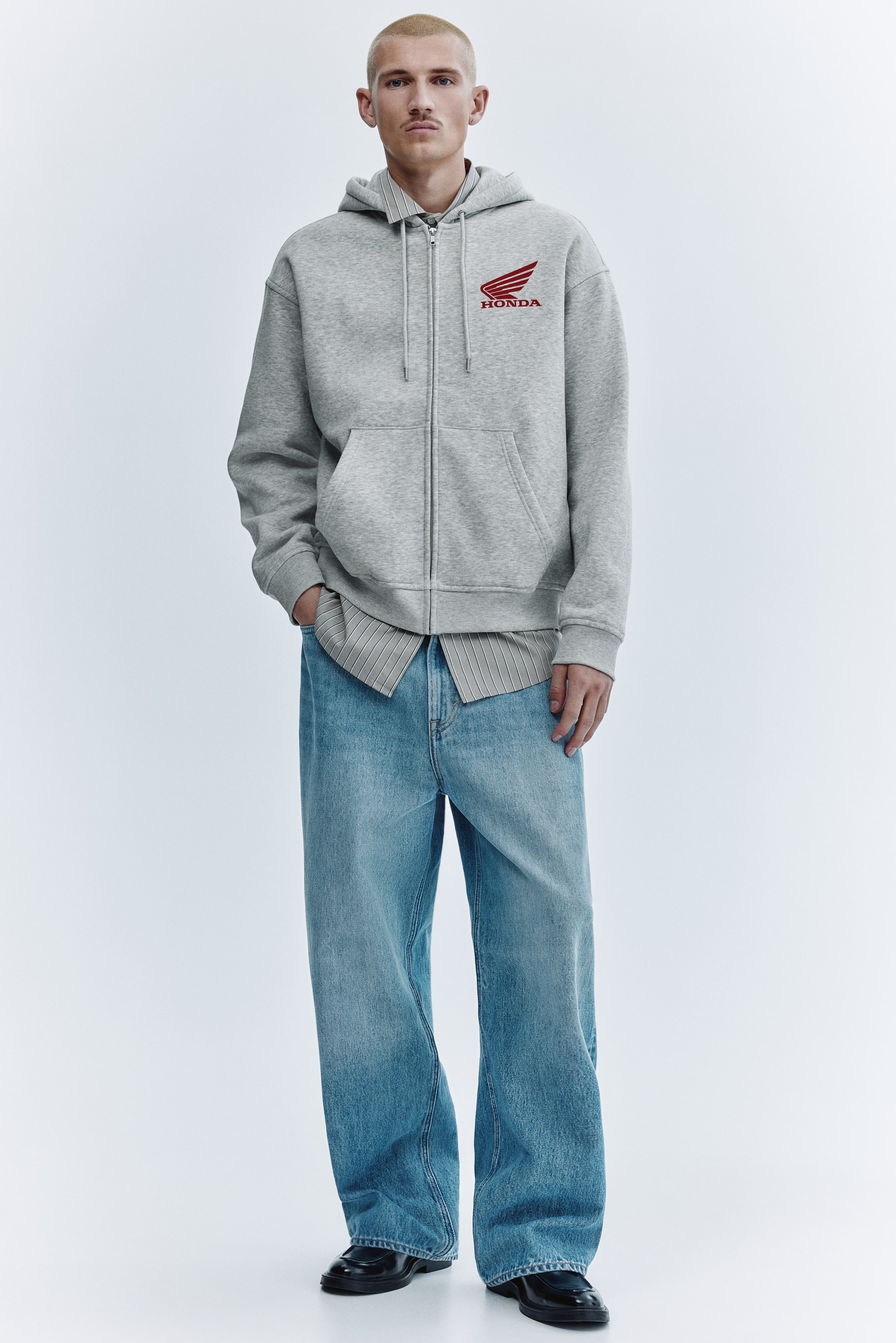 查看大圖: 這件灰色拉鍊連帽衛衣設有全長拉鍊、帶抽繩的連帽,以及位於左胸的紅色 Honda 標誌與翼形圖案。可見兩個前袋,分別位於拉鍊兩側,羅紋袖口與羅紋下擺完整了整體設計,衛衣套穿於一件淺色條紋有領襯衫外。