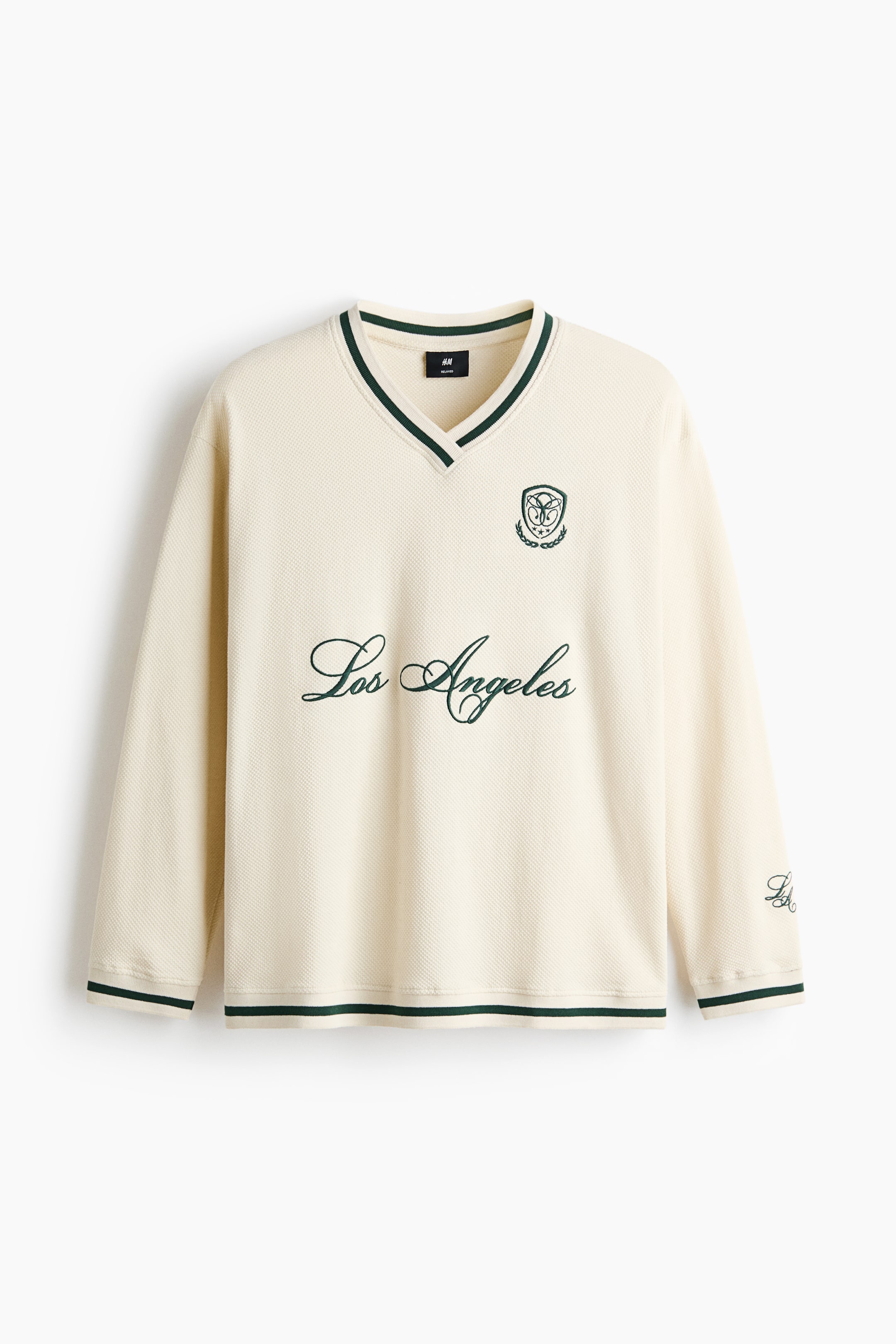 View larger image: A cream-coloured, long-sleeved V-neck shirt is shown from the front, displaying a textured knit fabric. It features dark green striped trim along the collar, cuffs, and hem, with a cursive "Los Angeles" script printed across the chest and a small crest logo on the left chest.