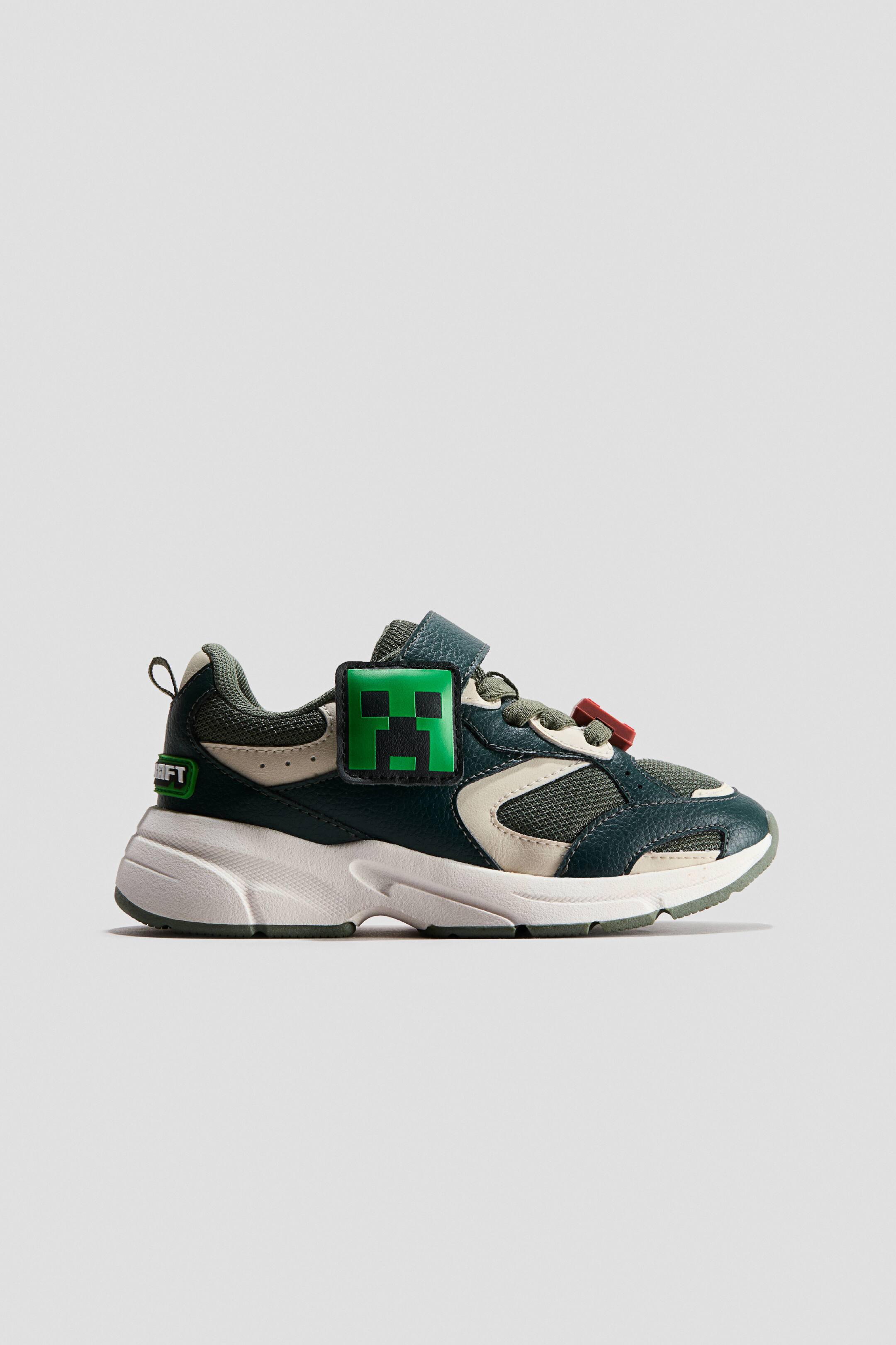 View larger image: These design-detail sneakers feature a paneled upper in dark green and beige with mesh inserts, presented in a side profile facing right. The sneakers display a chunky white textured midsole, a hook-and-loop strap closure, and a square green badge with a Minecraft Creeper design on the side.