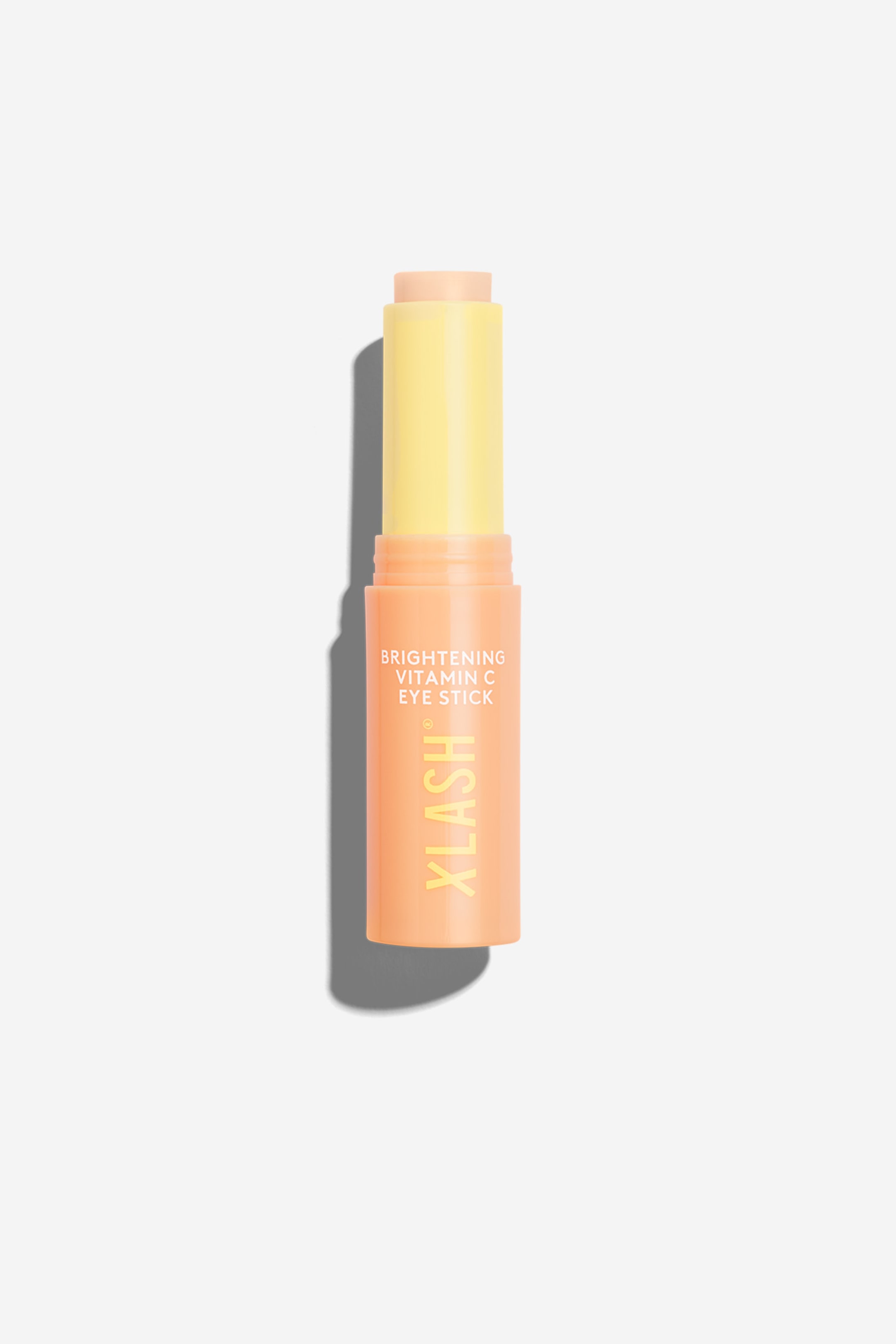 Vis større bilde: En sylindrisk øyestift står oppreist og vender fremover, med en lys ferskenfarget nedre del med gul tekst som lyder 'BRIGHTENING VITAMIN C EYE STICK' og 'XLASH'. Den gule øvre hetten avslører en lys ferskenfarget produktstift som stikker ut fra den.
