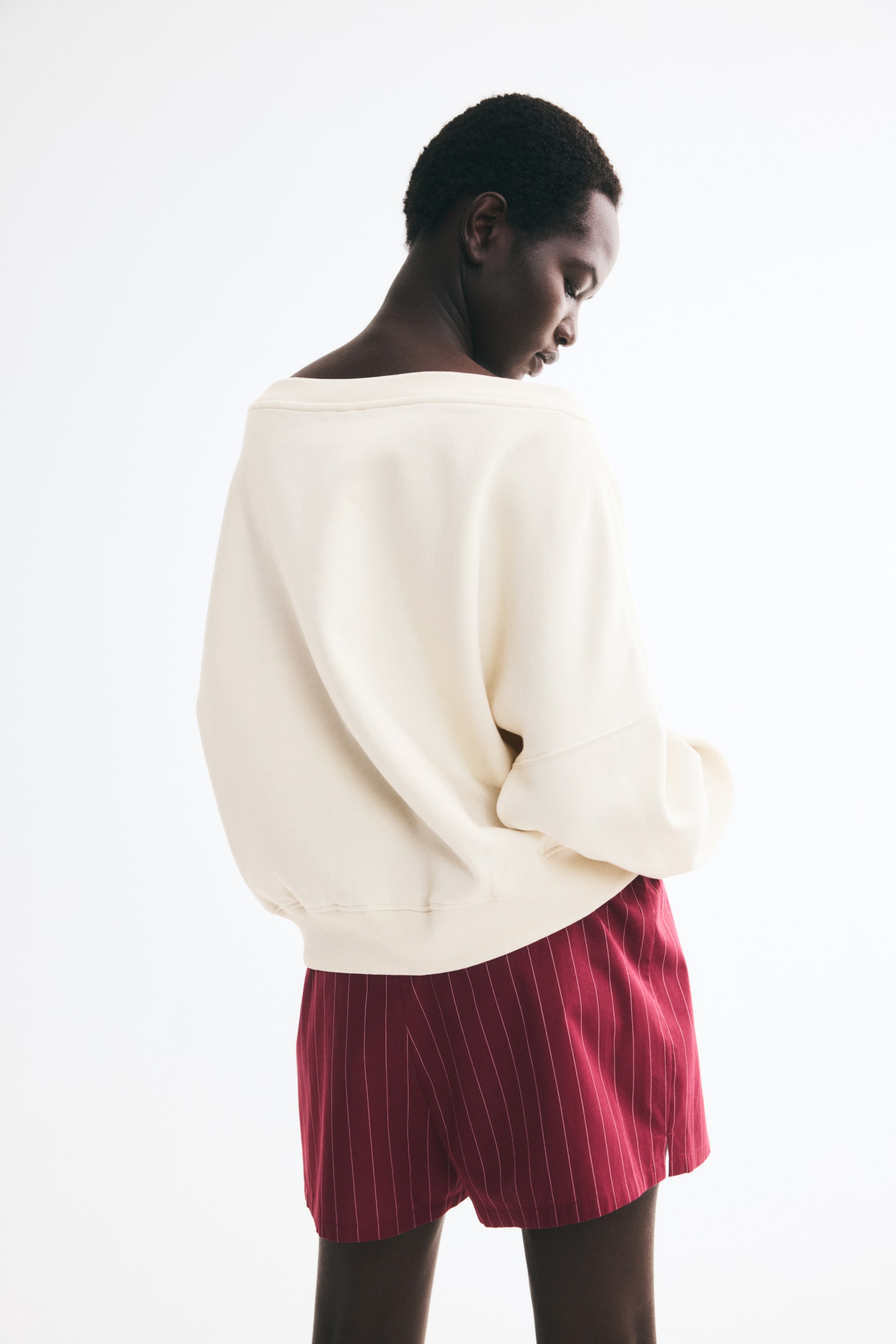 View larger image: A cream-coloured, off-the-shoulder sweatshirt is being worn by the model, shown from a three-quarter back view. The relaxed-fit sweatshirt features a wide neckline, dropped shoulders, and a ribbed hem, and is styled with red pinstriped shorts.